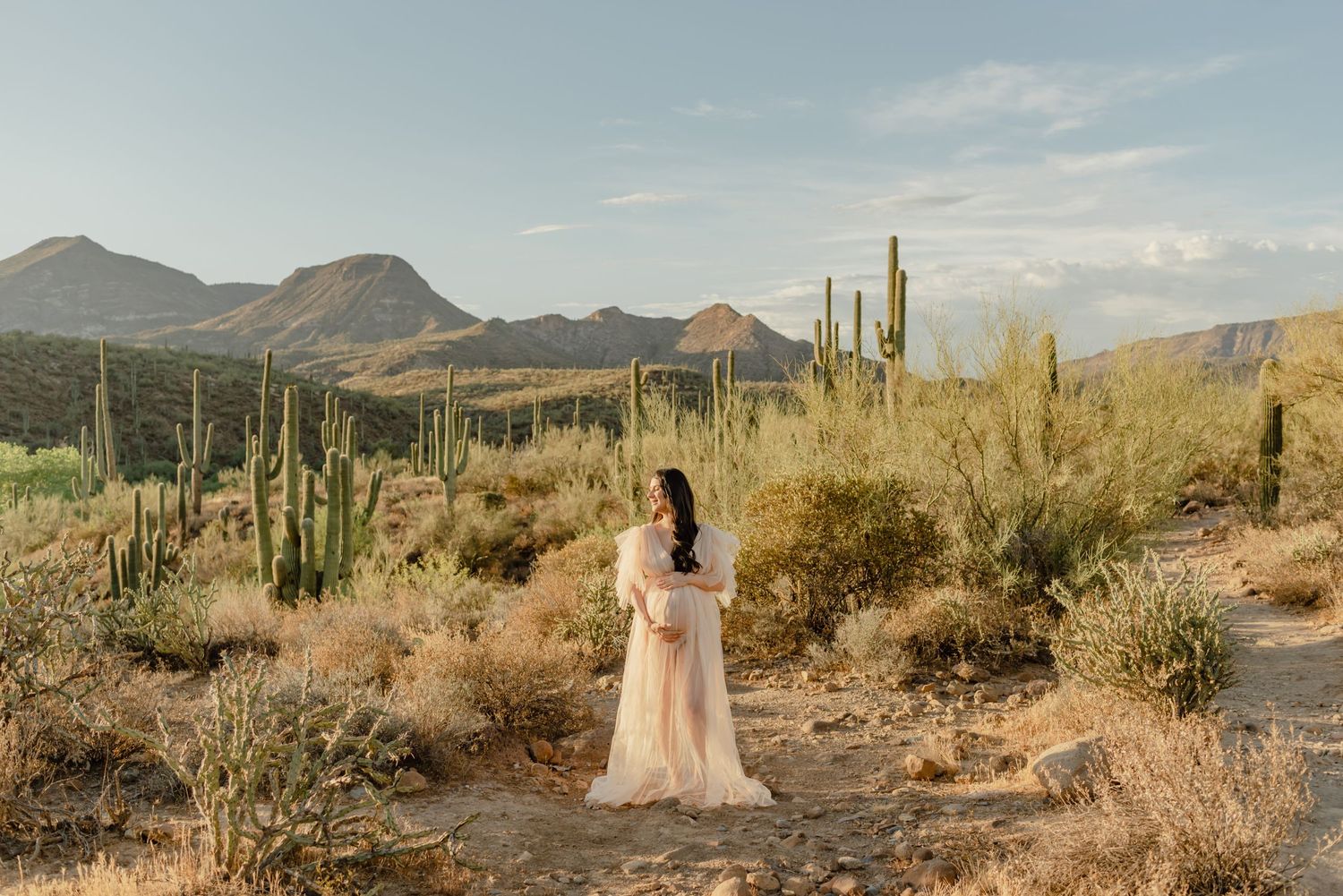 pregnant woman in a flowing gown smiles among saguaro cacti at golden hour, phoenix desert maternity photography session
