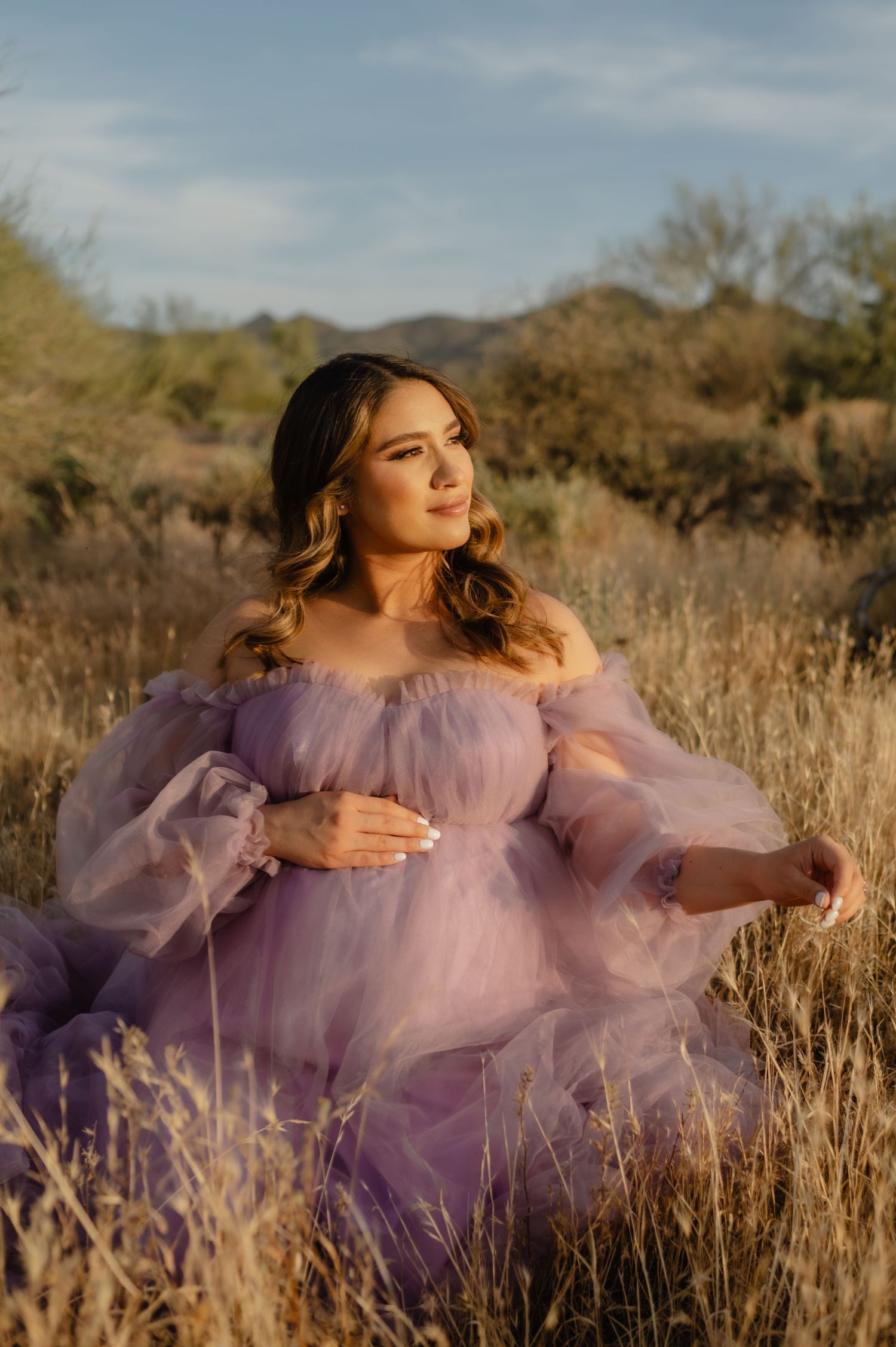 Pregnant woman in lavender tulle off-shoulder gown sitting in golden dry grass field at sunset.