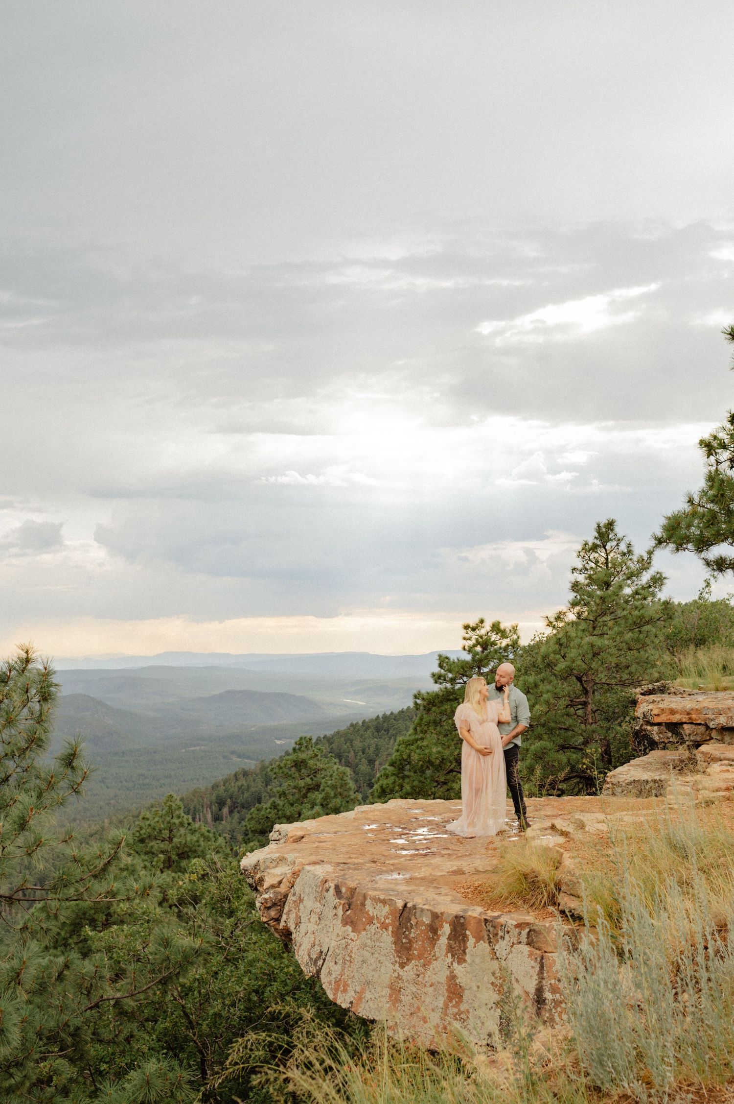 Couple embracing on a rocky cliff overlook surrounded by pine trees with dramatic cloudy sky at sunset.