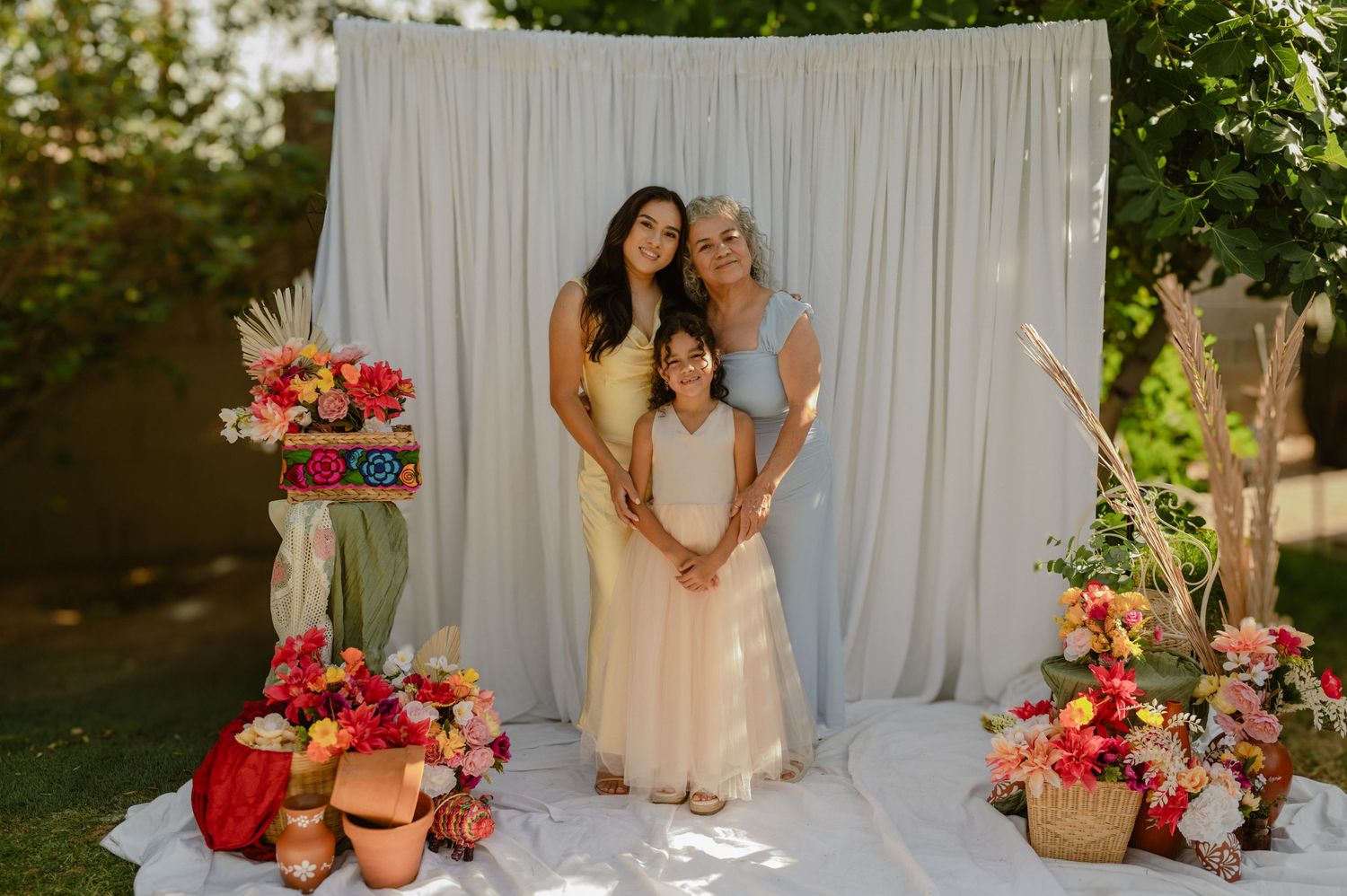three generations of women embrace in front of a white draped backdrop surrounded by fresh flowers, glendale mini session