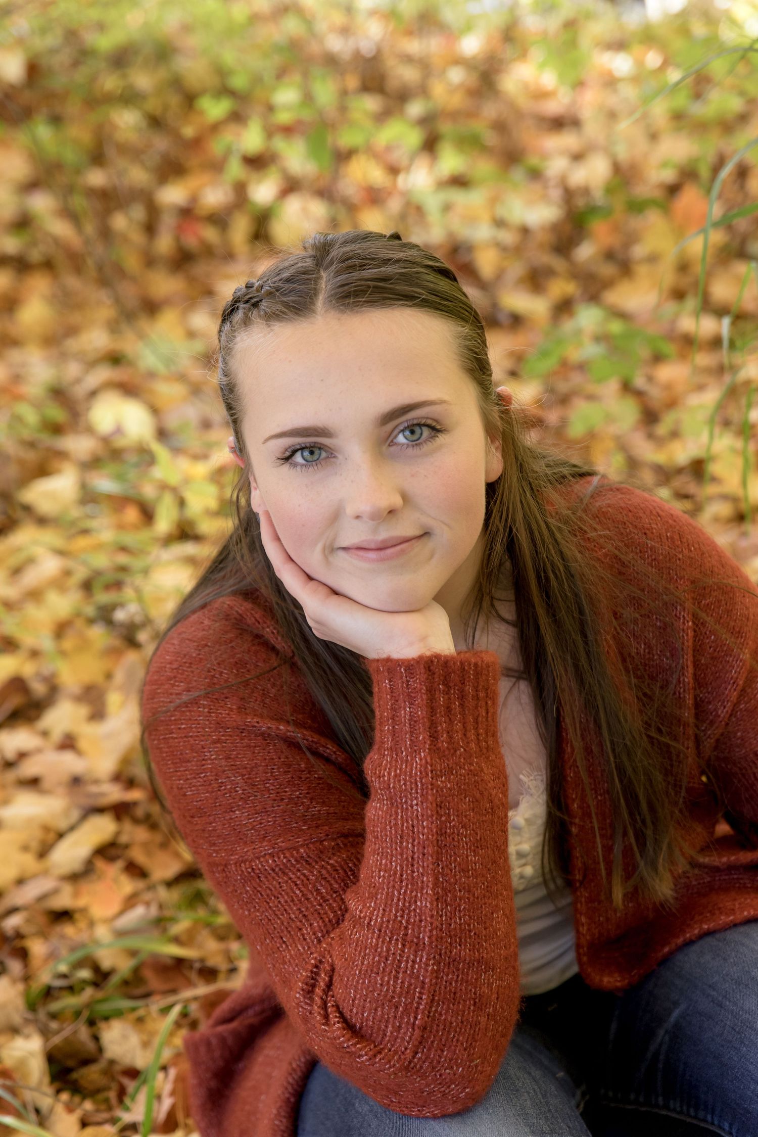 Teen girl with braided hair rests chin on hand sitting among fallen autumn leaves in rust cardigan. West Alton Marina Alton NH