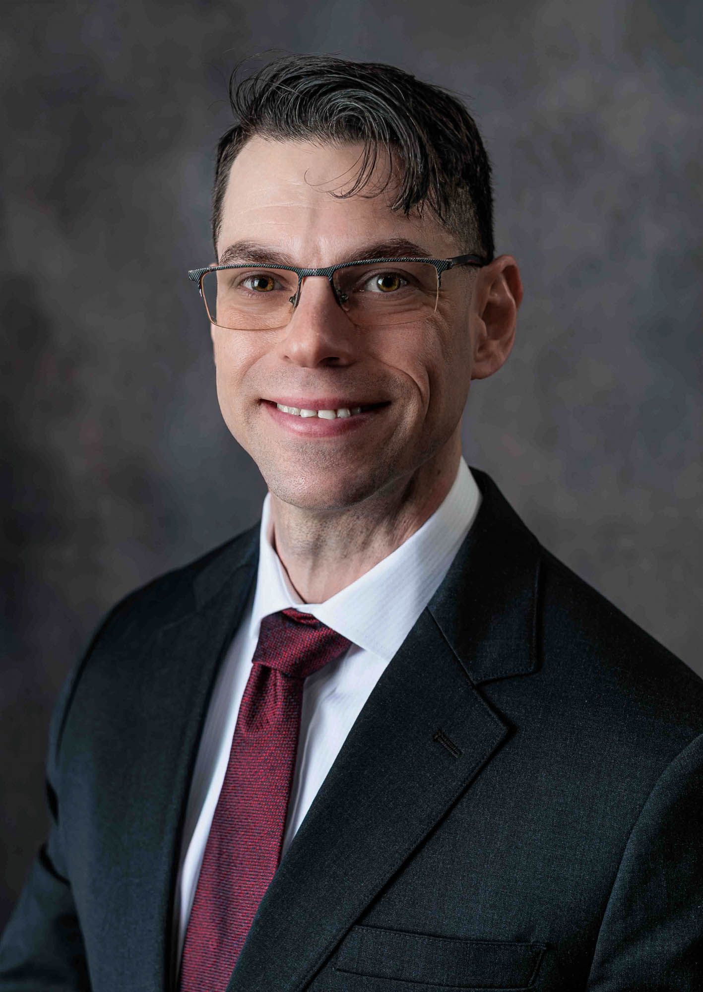 Professional headshot of a man from Concord NH in a dark suit and red tie wearing glasses, smiling confidently at our studio in Barnstead.