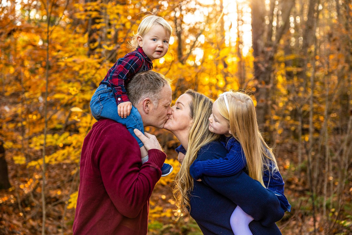 Parents kissing while holding two young children in a vibrant autumn forest with golden fall foliage. Cotton Valley Rail Trail Wolfeboro NH