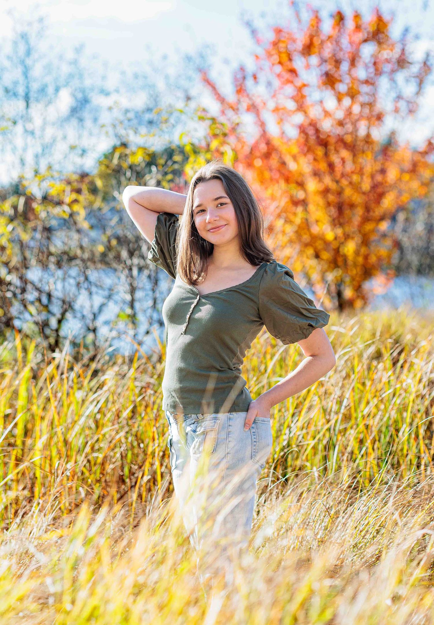Young woman in olive green top stands in tall golden grass with colorful autumn foliage behind her. Locke Lake Barnstead NH