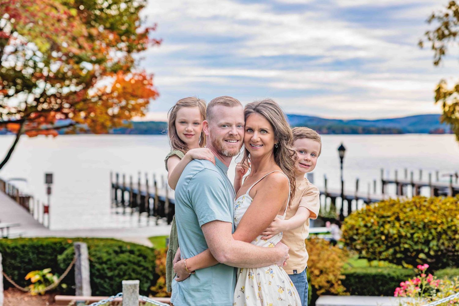 Smiling family of four with two young children embracing outdoors near a lake with fall foliage backdrop. Cate Park Wolfeboro NH