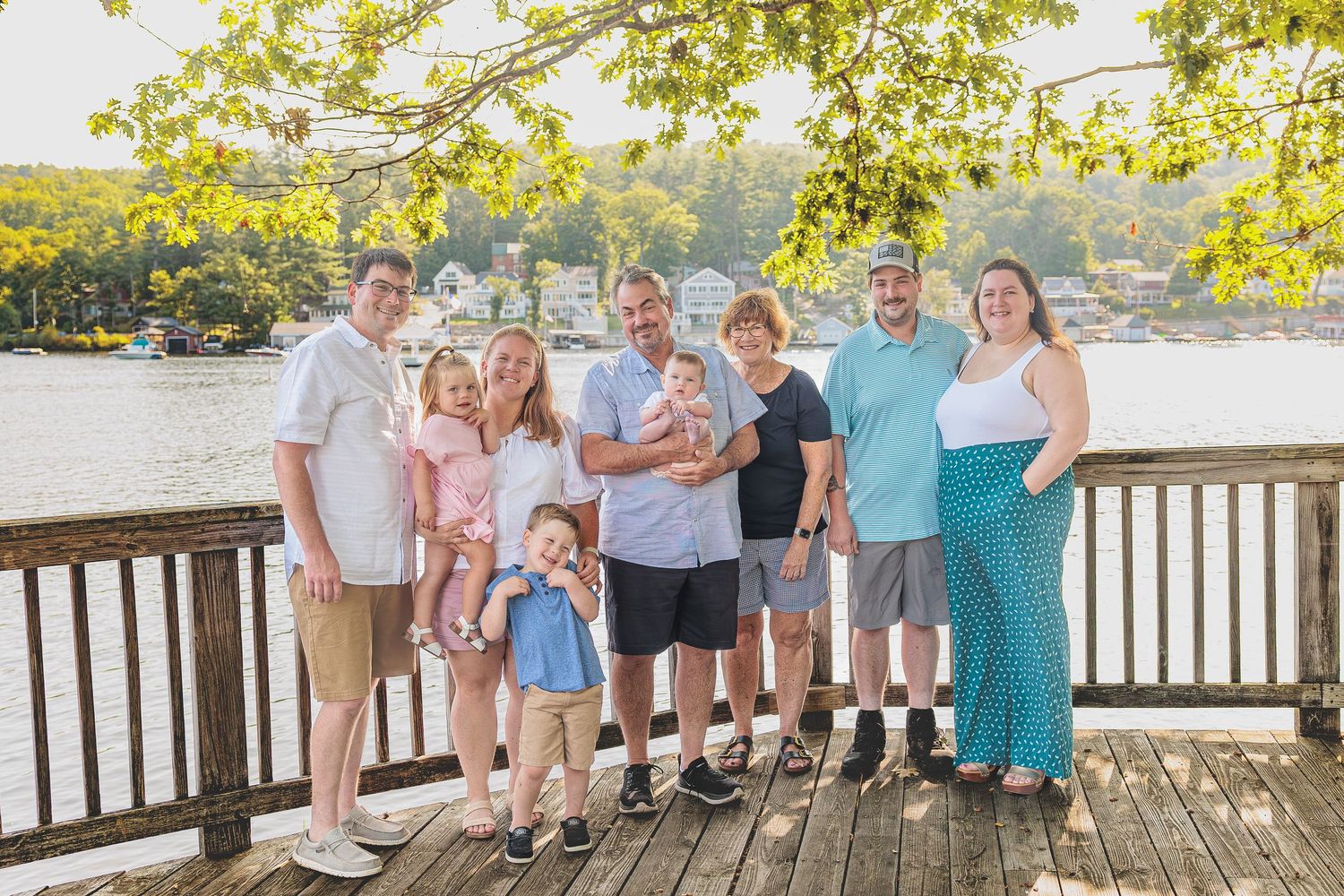 Extended family group of eight posing on a wooden dock with a scenic waterfront village in background. Alton Bay NH