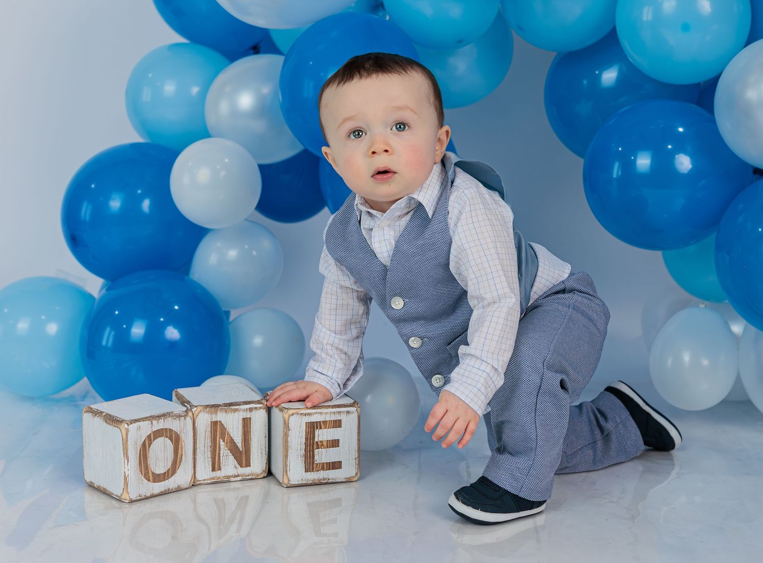 Baby boy dressed in a vest sitting with wooden ONE blocks at his first birthday party.