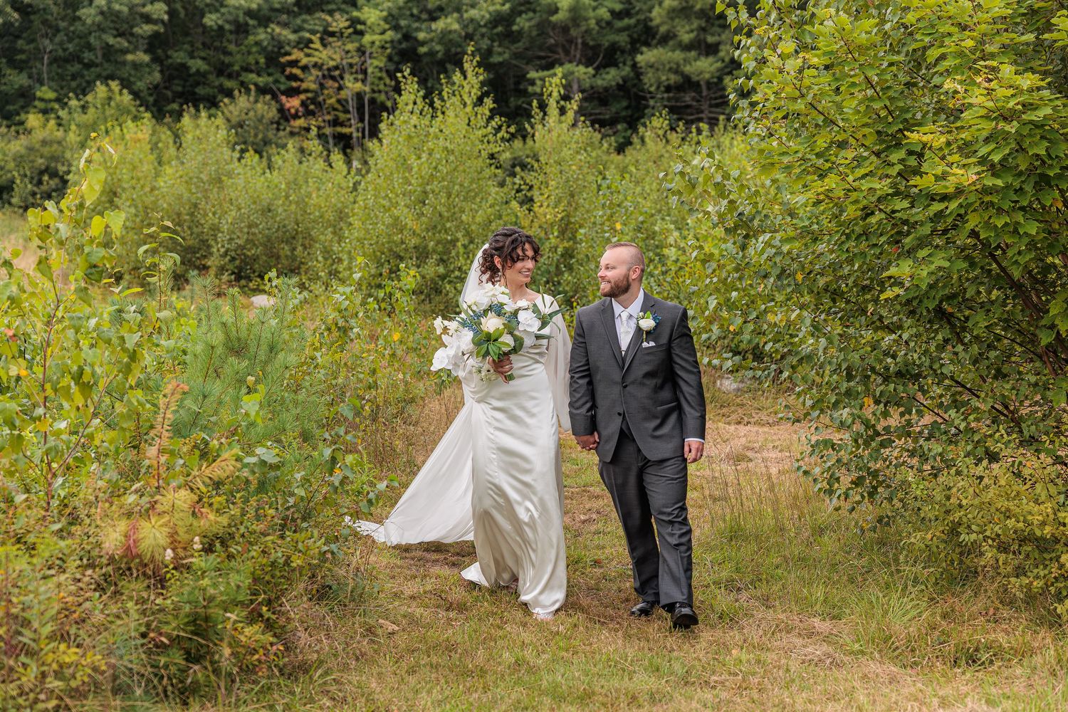 Bride and groom walk hand in hand along wooded path through lush summer greenery. The Venue at Ponderosa Milton NH