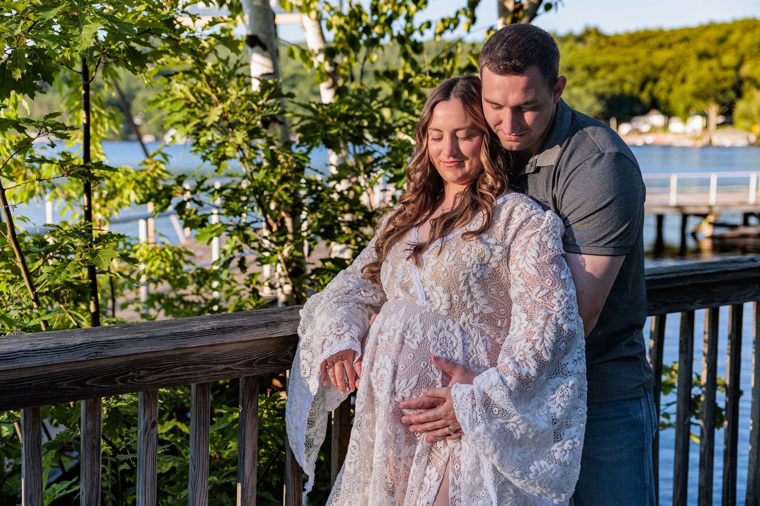 A couple tenderly embraces on a waterfront deck at sunset, cradling a baby bump in a lace gown. Alton Bay NH