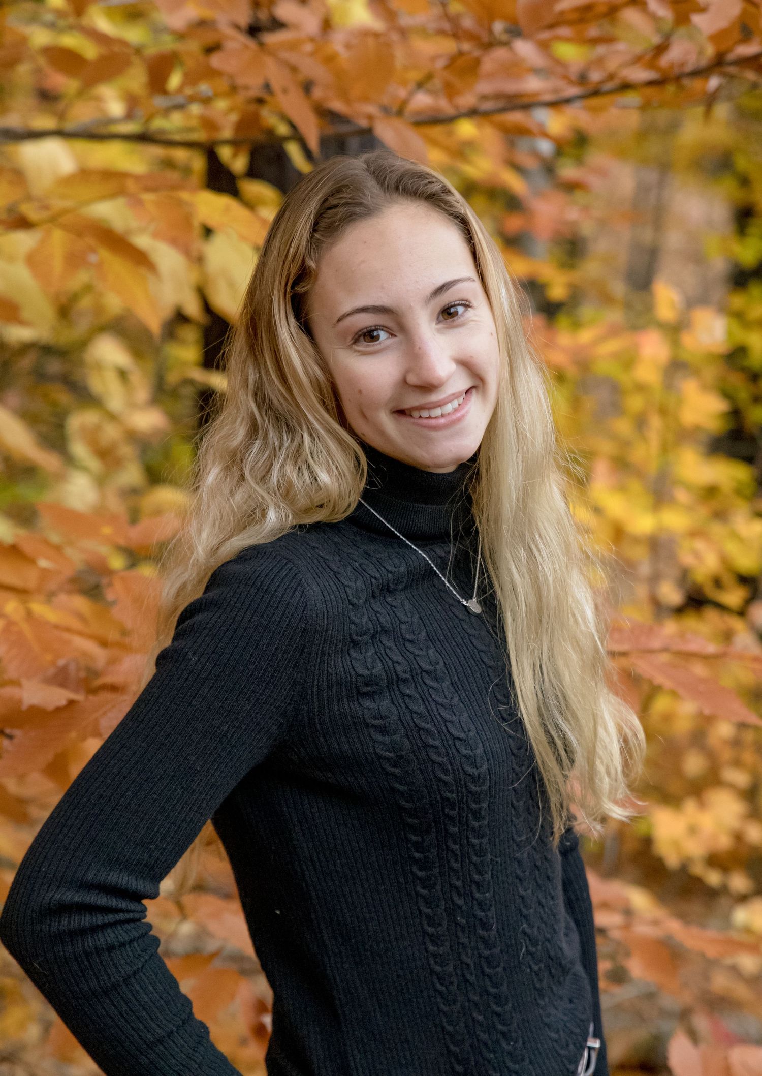 Blonde teenage girl smiles over her shoulder wearing black turtleneck sweater with fall foliage background.