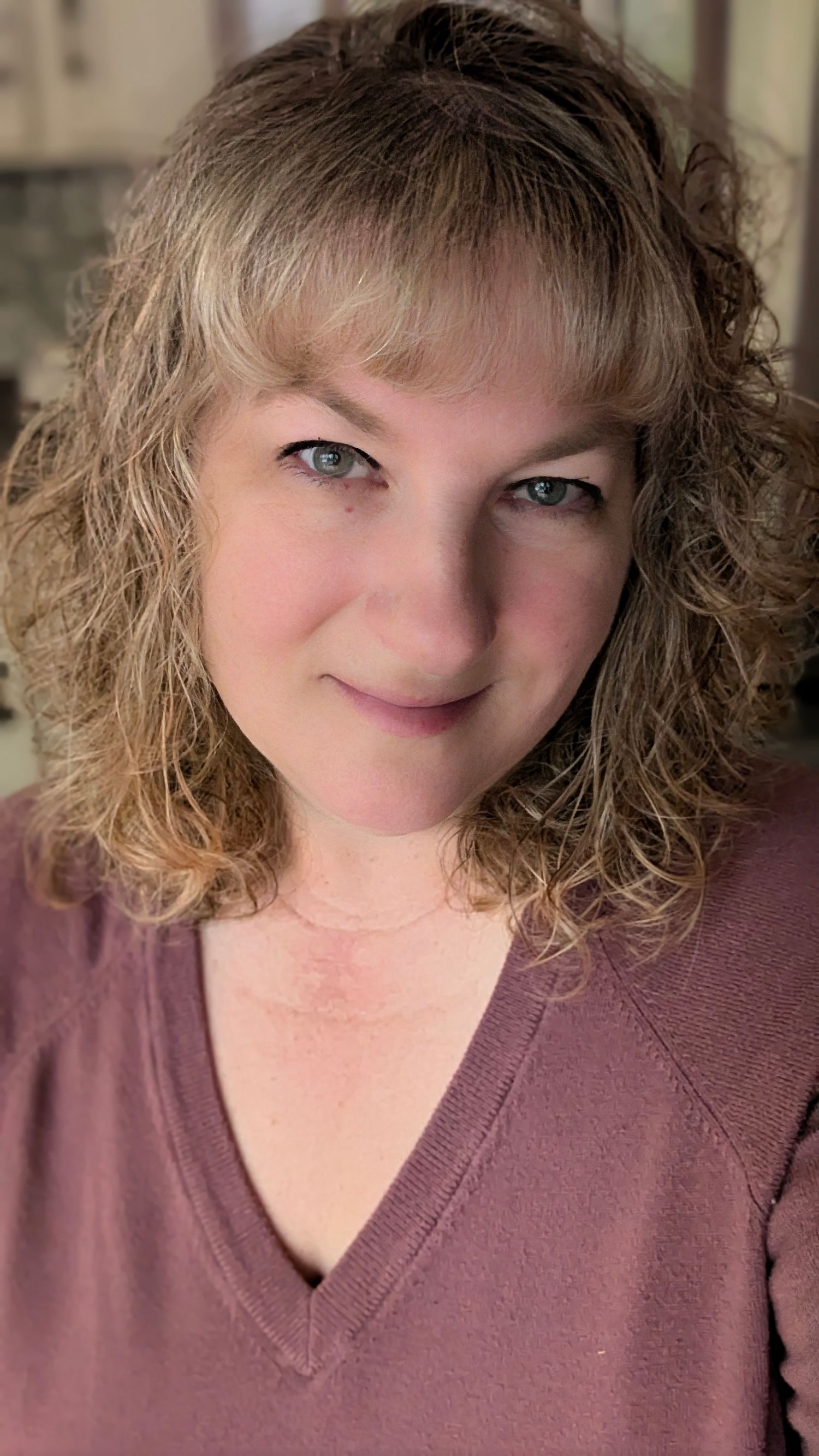 A woman with curly blonde hair and blue eyes smiles softly, wearing a mauve v-neck sweater.