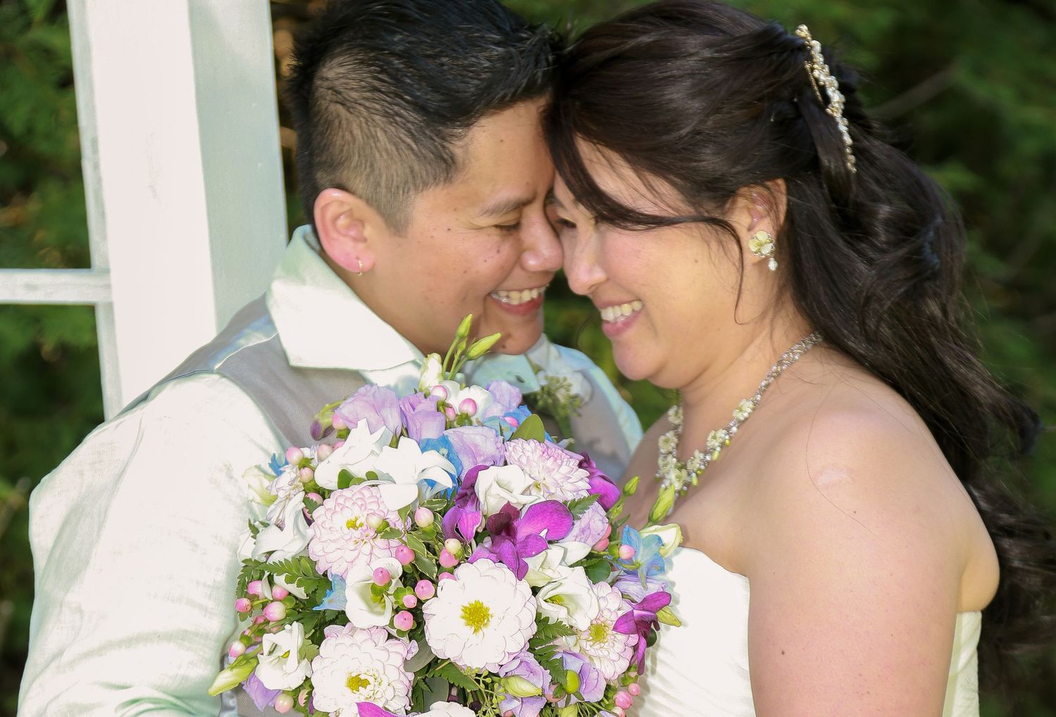 Bride and groom touching foreheads romantically, bride holding colorful bouquet of pink and purple flowers at outdoor wedding.