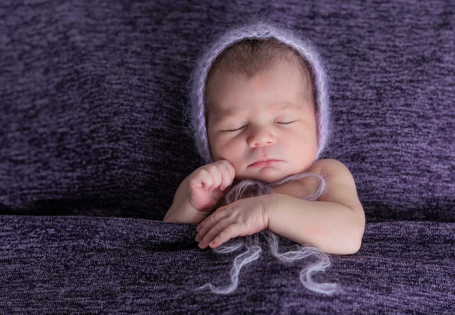 Newborn baby sleeping peacefully on purple fabric, wearing a soft lavender knit bonnet, with tiny hands folded.