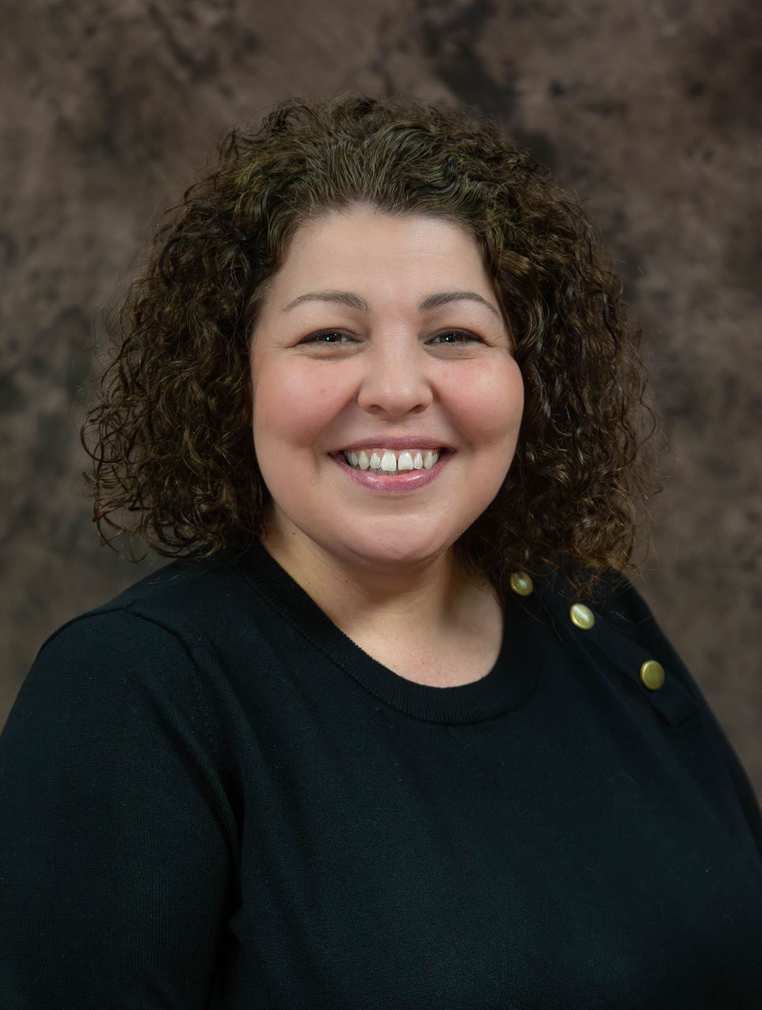 Cheerful curly-haired woman smiling broadly in black top against brown studio backdrop.
