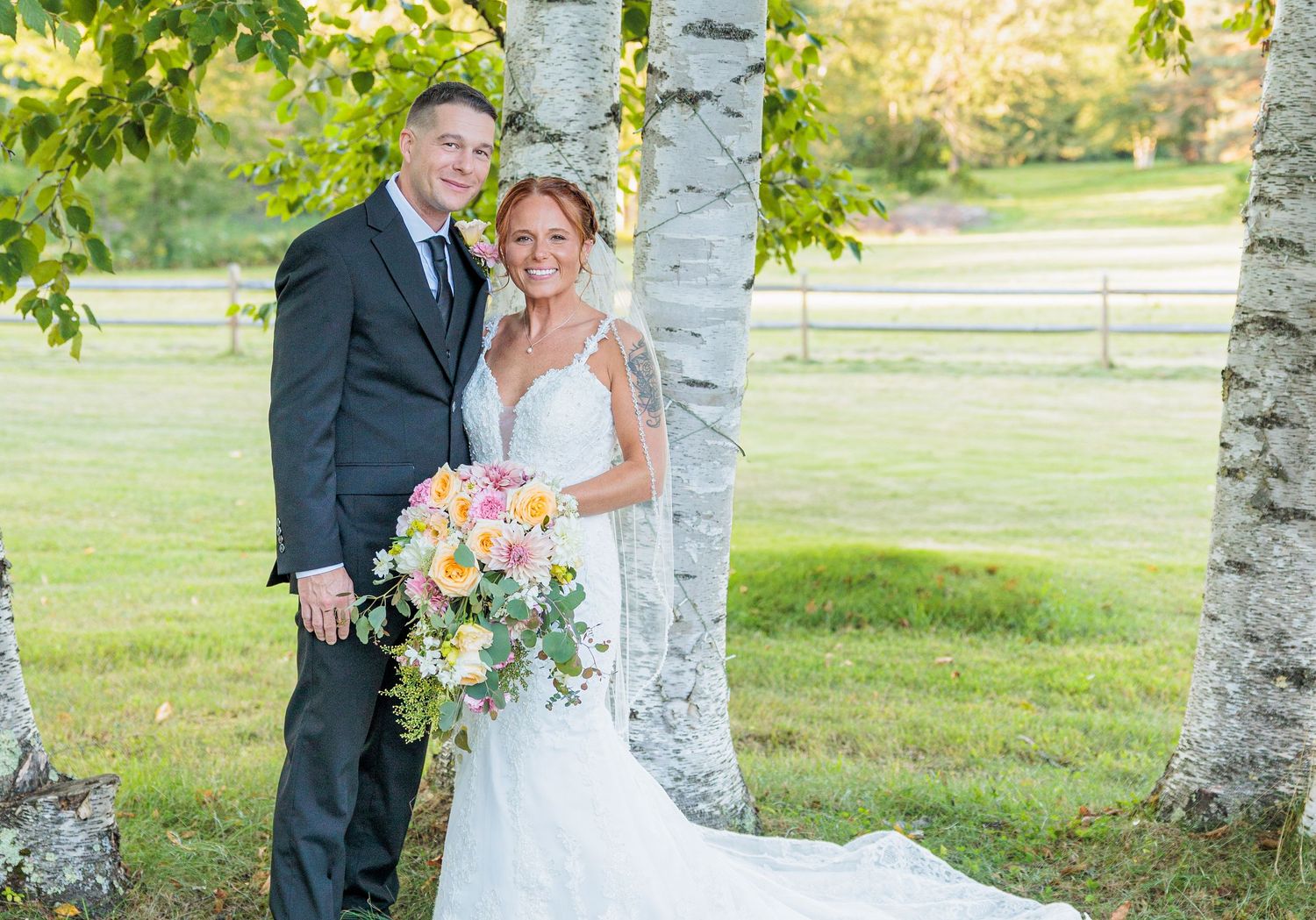 Couple poses in wedding attire beside white birch trees holding colorful peach and yellow bouquet. Top of the Ridge Farm New Durham NH