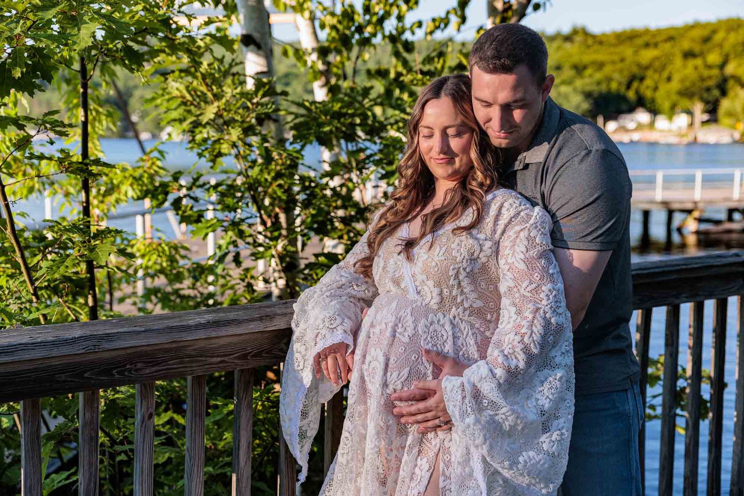 Couple sharing a tender maternity moment on a wooden deck surrounded by lush greenery near a waterfront at sunset. Alton Bay NH