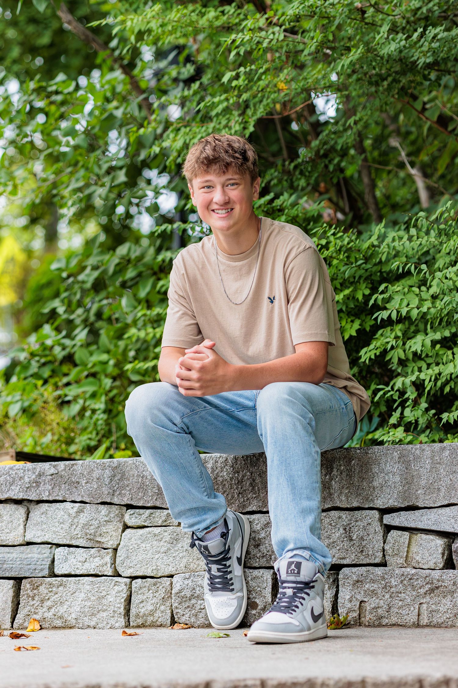 Smiling teenage boy sitting on stone wall outdoors surrounded by lush green trees. Cate Park Wolfeboro NH