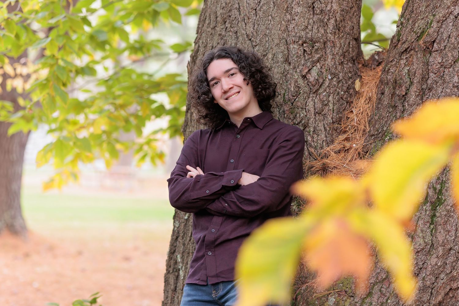 Young man with curly hair leans against a tree with arms crossed during autumn outdoor portrait session. Bear Brook State Park Suncook NH