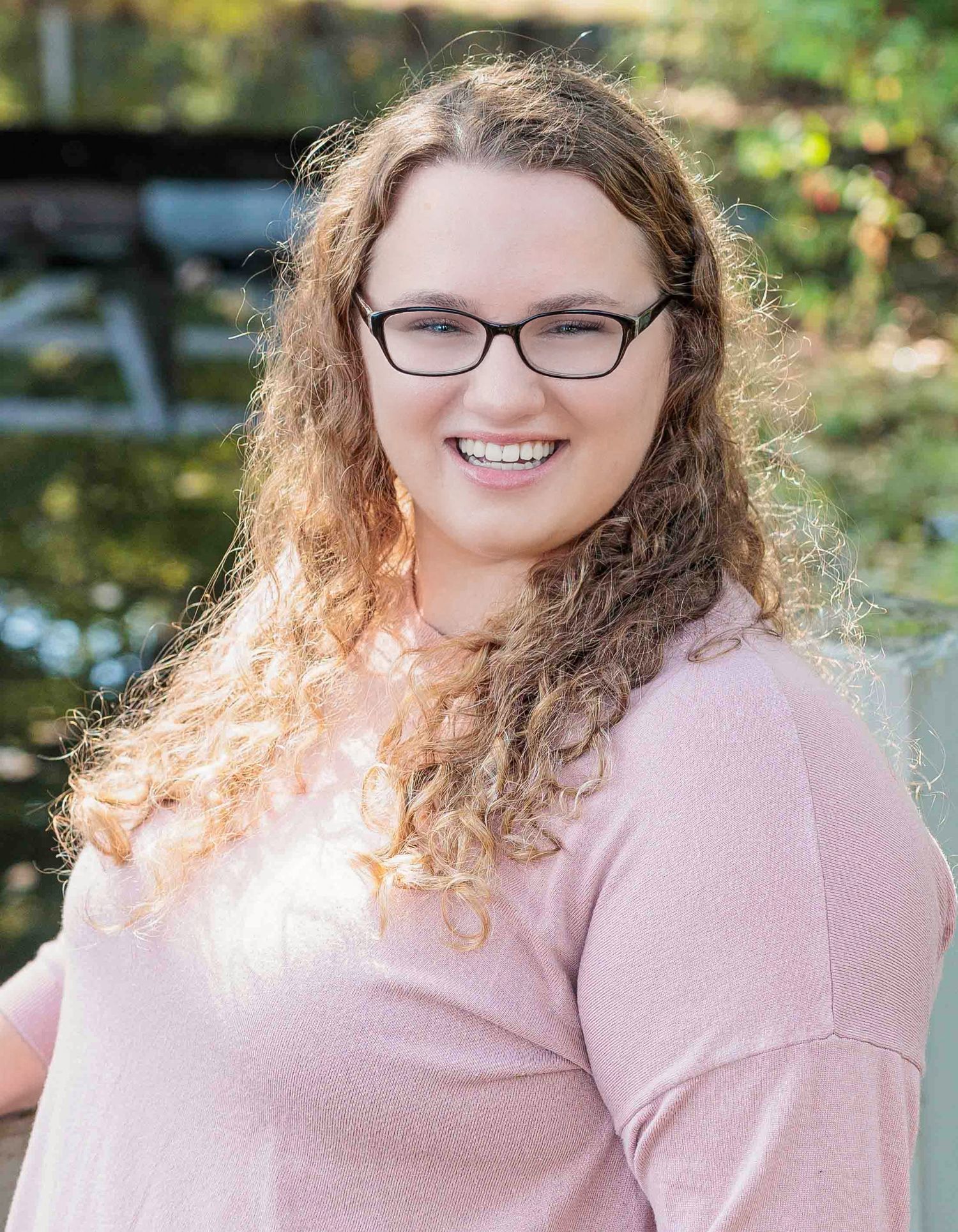 Young woman with curly hair and glasses smiling outdoors in pink sweater. Bear Brook State Park Suncook NH