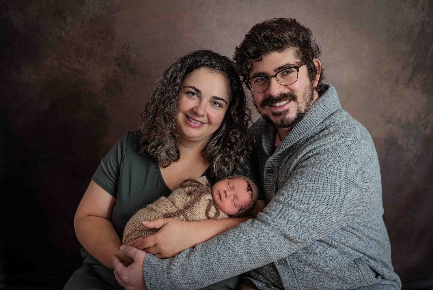 Smiling new parents hold their sleeping newborn baby together against a warm studio backdrop.