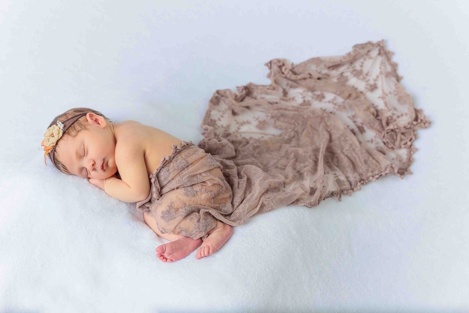 Sleeping newborn baby wrapped in soft taupe lace blanket with floral headband on white backdrop.