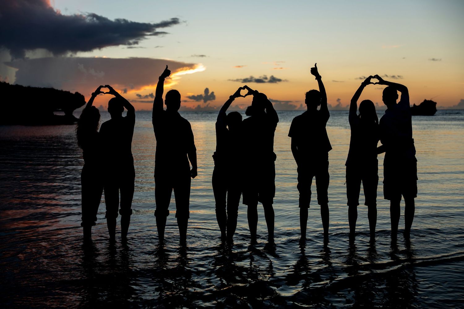 Silhouetted figures stand along a beach at sunset making heart shapes with their arms against an orange sky.