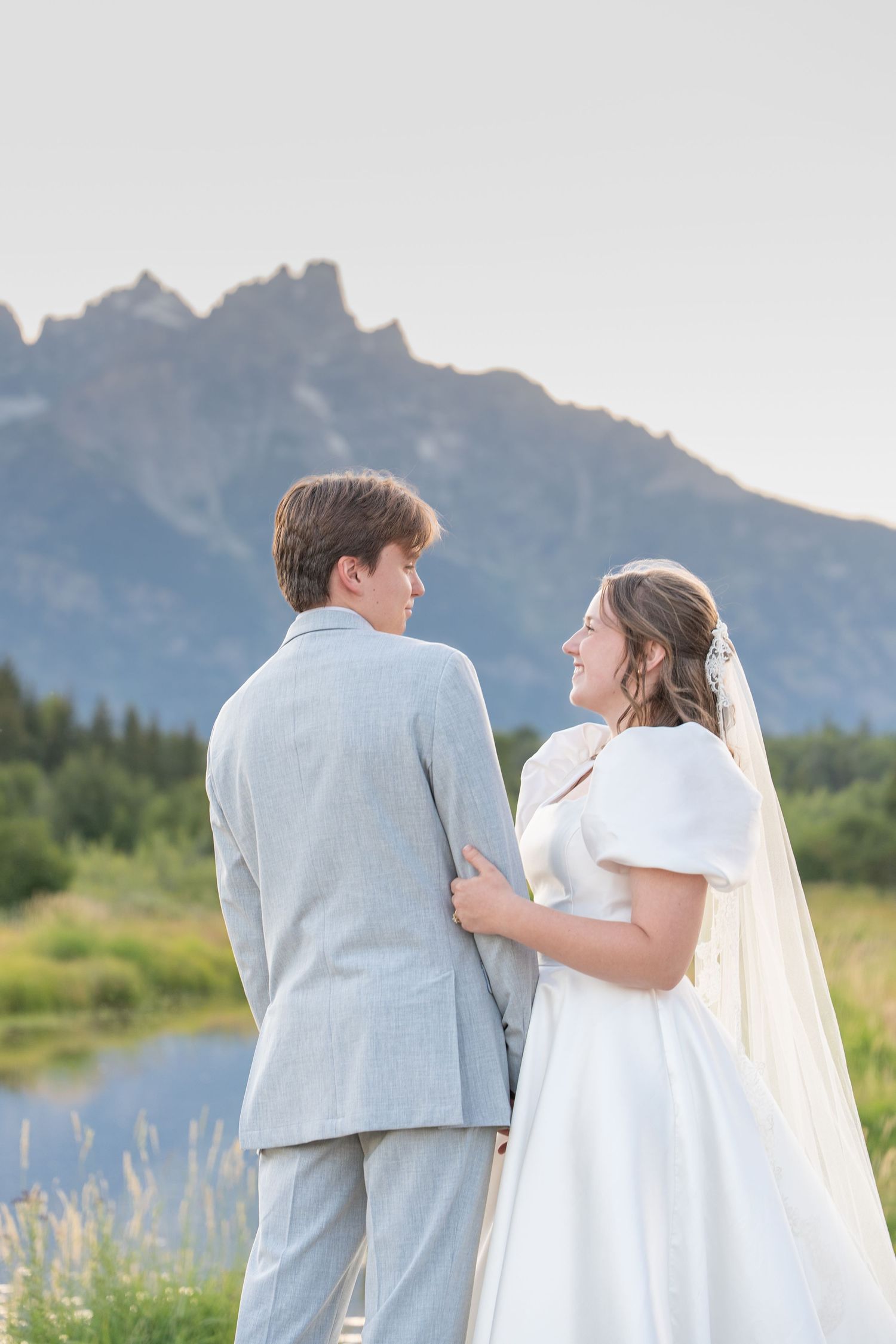 A couple in wedding attire share a tender moment against a backdrop of majestic mountains and a serene lake.