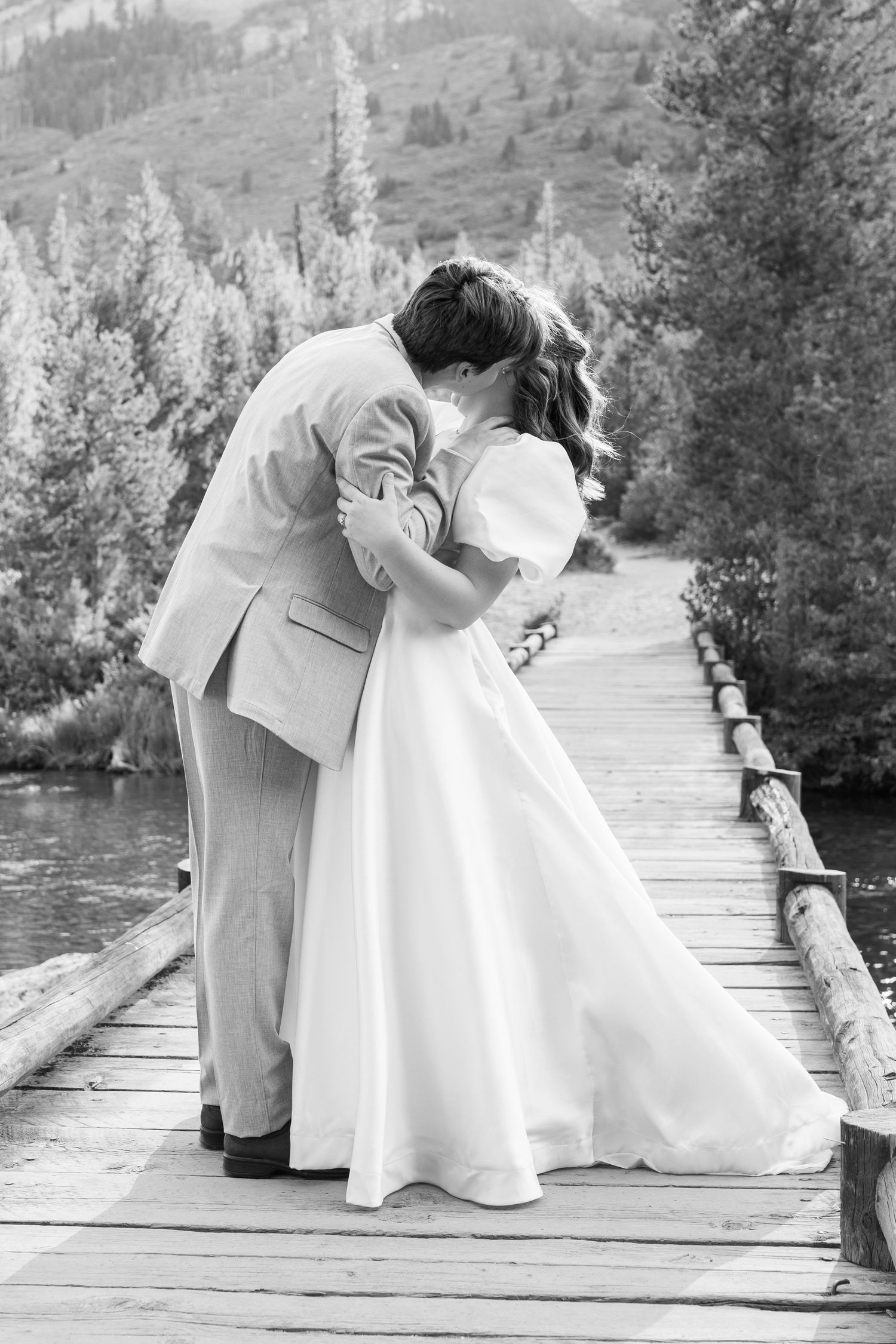 A romantic black and white wedding photo of a couple embracing on a wooden bridge in the mountains.