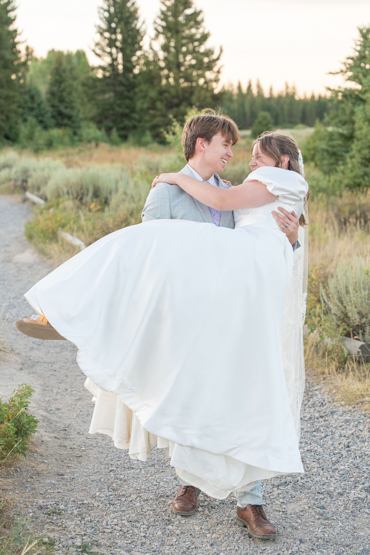 Groom carries bride in white dress along a gravel path with evergreen trees in background.