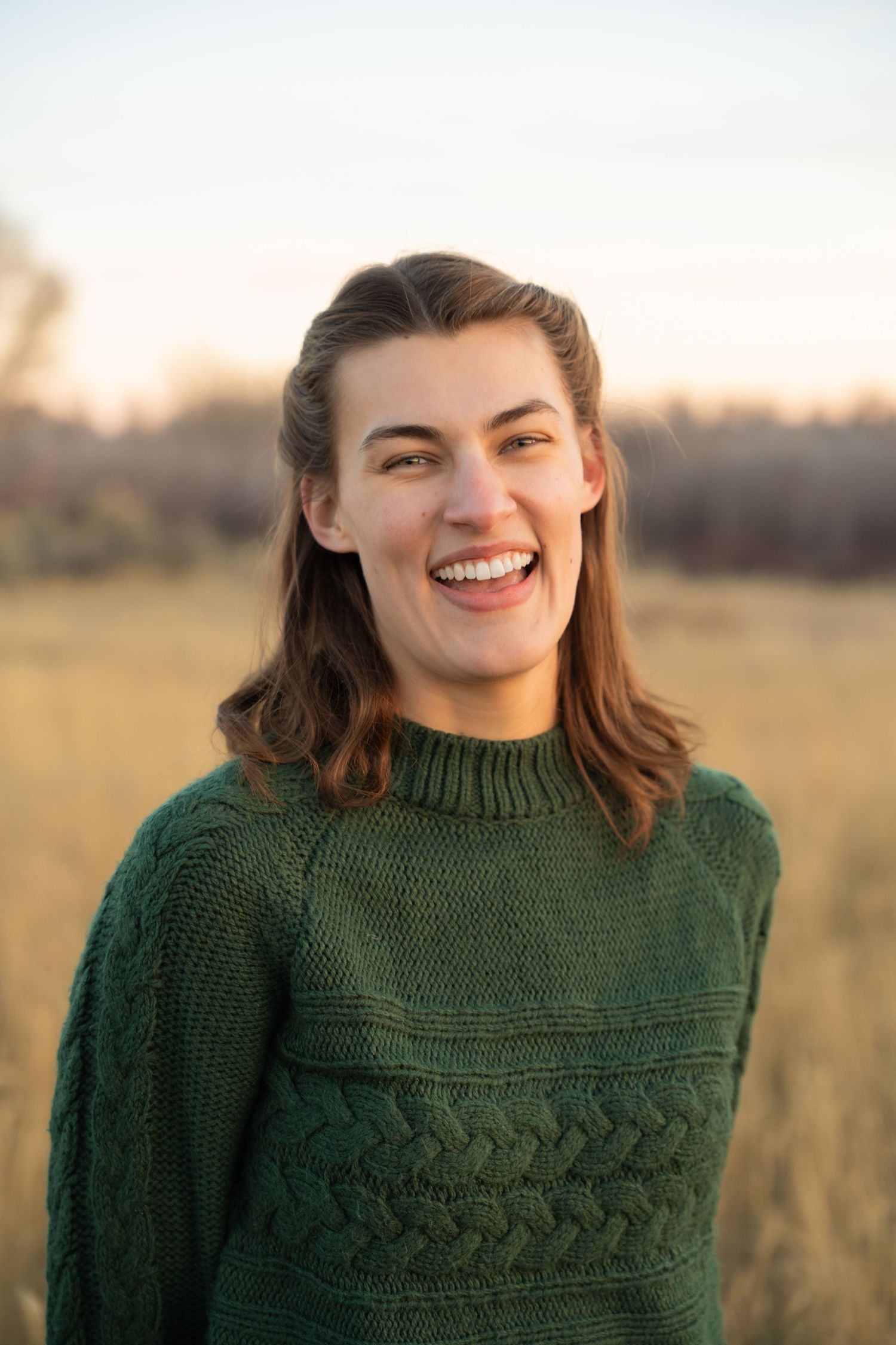 Person in a green cable knit sweater smiling in a sunny outdoor setting.