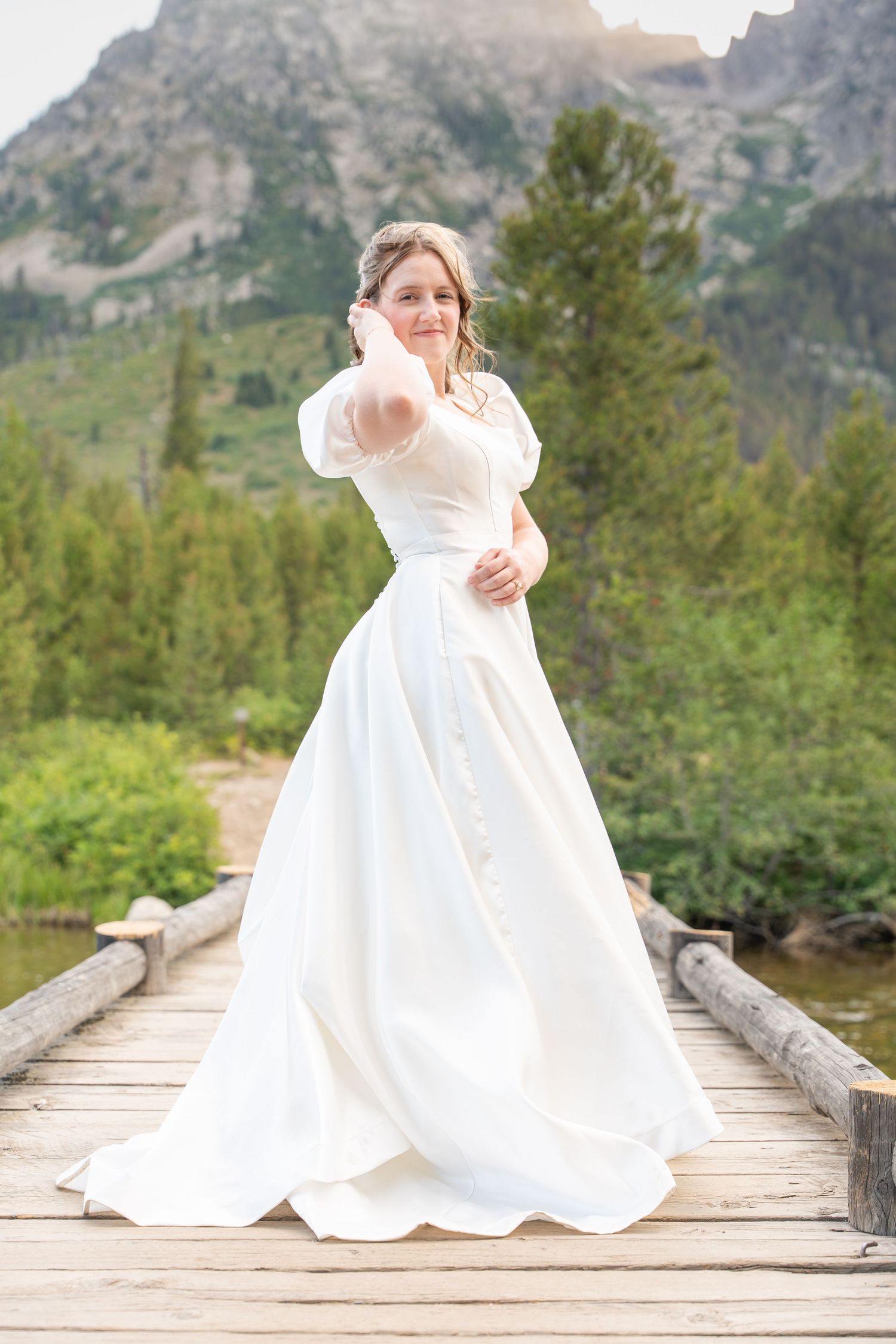 A bride in a classic white wedding dress poses on a rustic wooden bridge with mountains in the background.