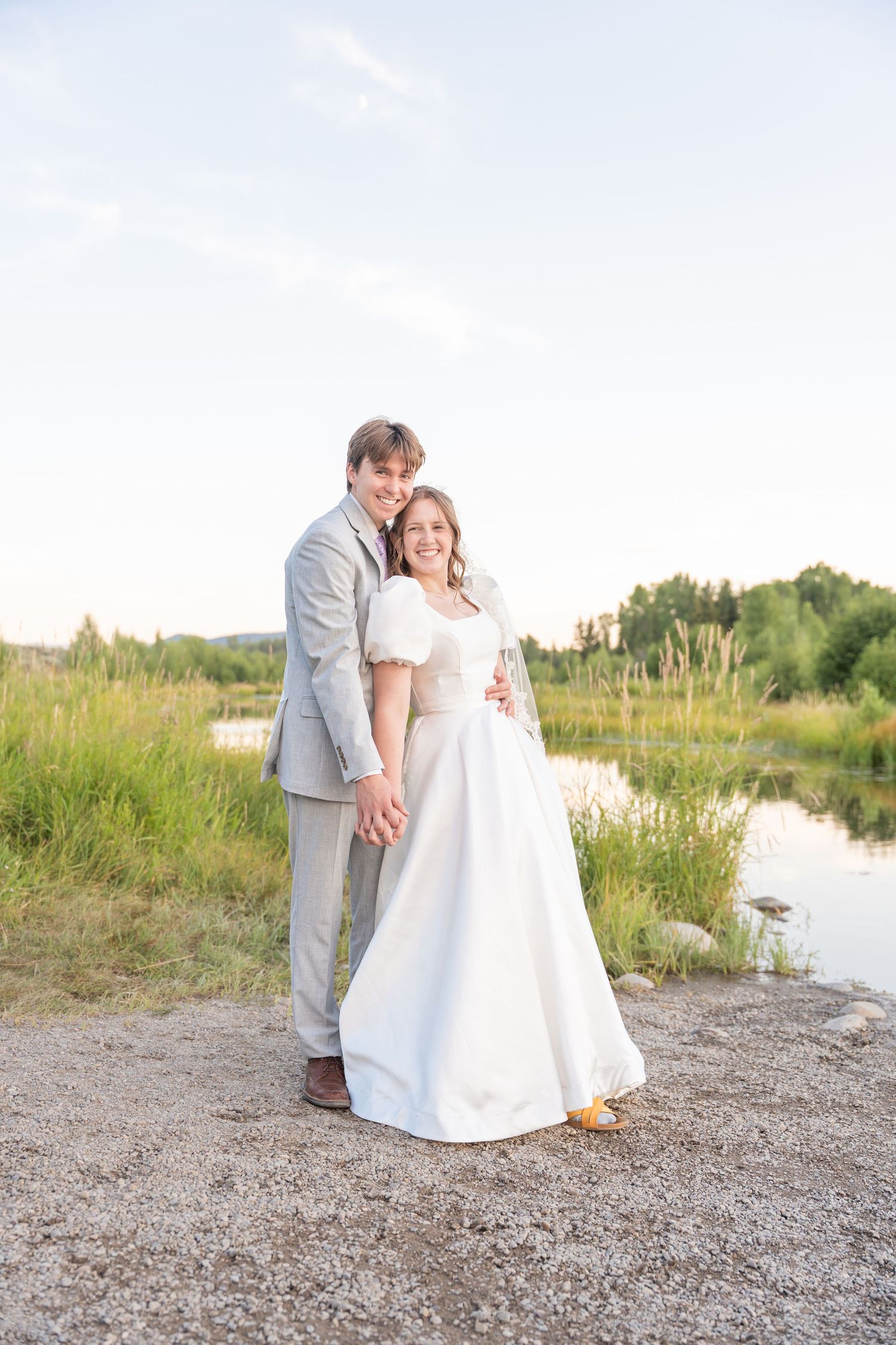 A newlywed couple poses together on a lakeside path with mountains and forest in the background.