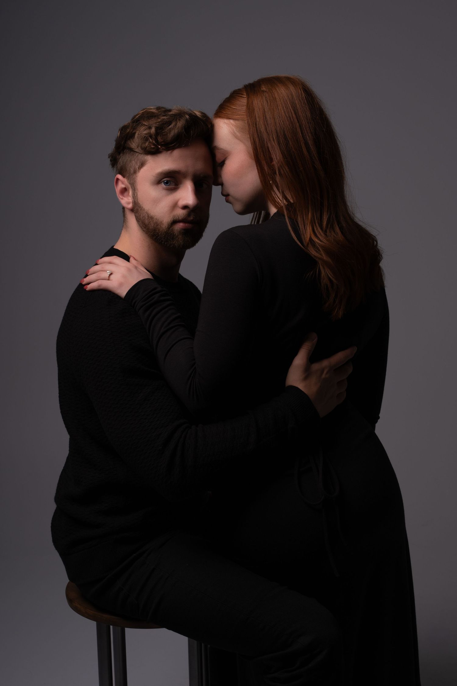 A couple in black clothing embrace intimately against a dark background in a moody studio portrait.