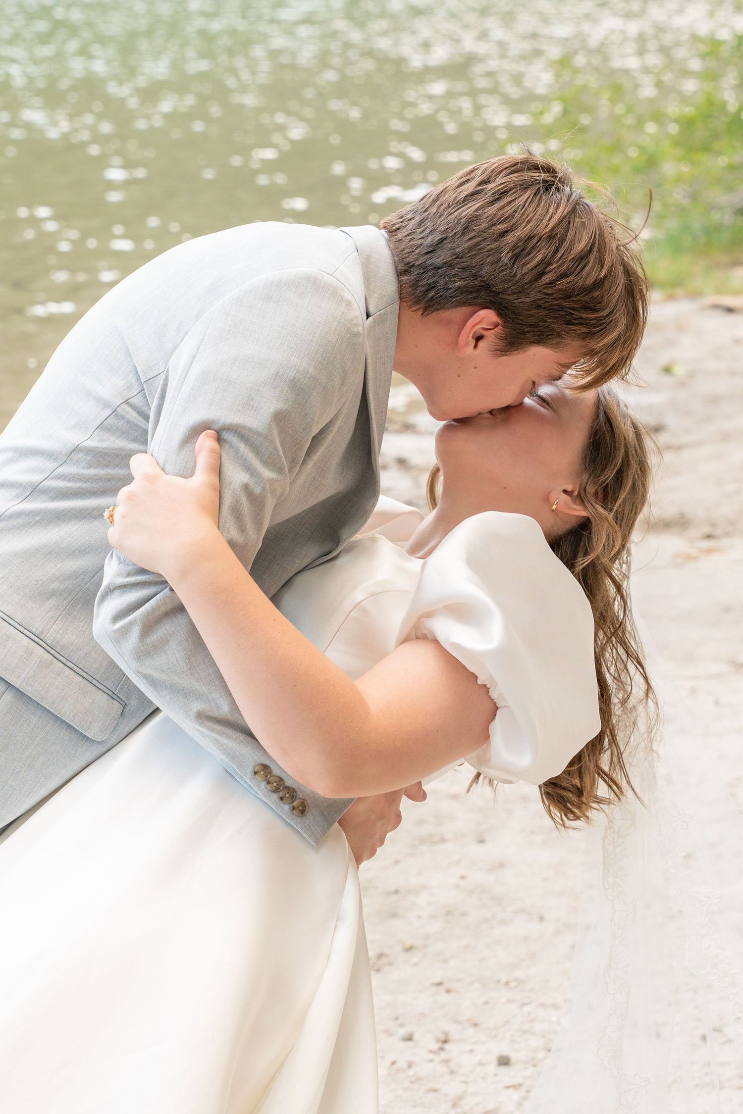 A romantic dip kiss between newlyweds by a serene lake on their wedding day.