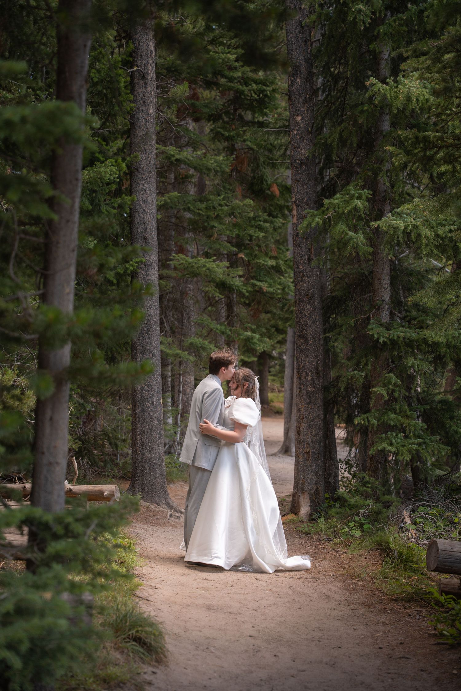 A romantic wedding portrait of a couple embracing on a forest path surrounded by tall pine trees.