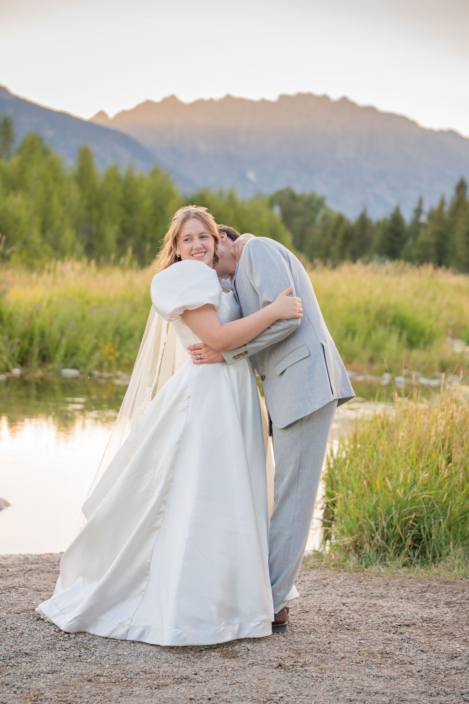A romantic embrace between newlyweds in a light gray suit and white dress near a mountain lake.