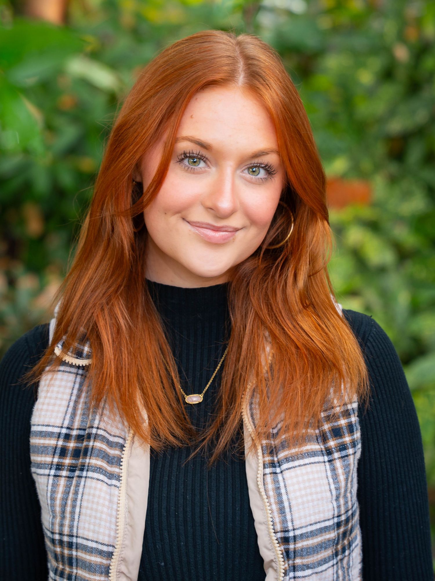 Portrait of someone in a black turtleneck and plaid vest with vibrant foliage background.