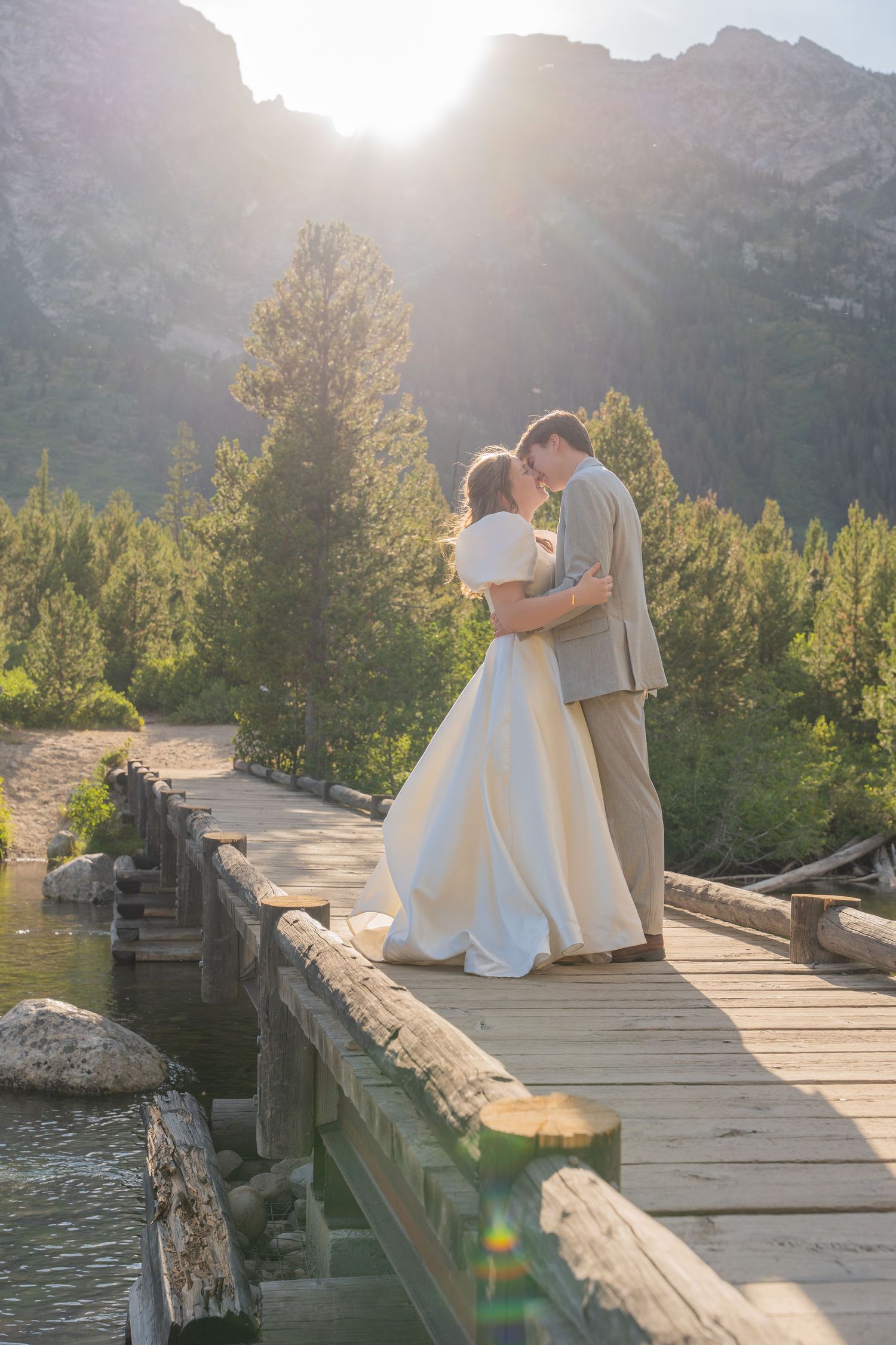 A wedding couple embraces on a wooden bridge at sunset with mountain peaks and pine trees in the background.