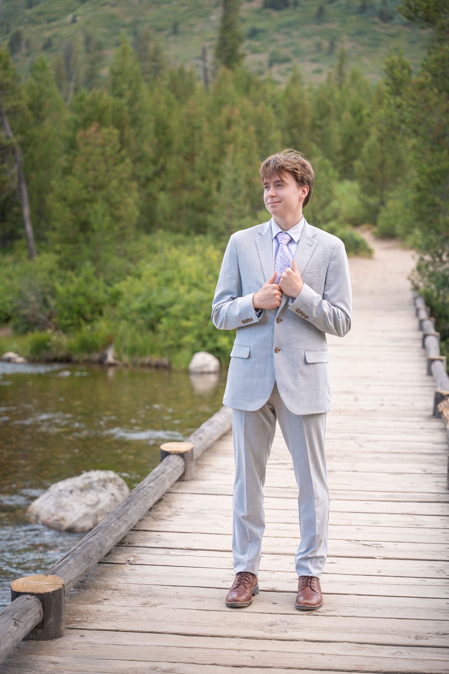A person in a light gray suit stands on a wooden boardwalk surrounded by mountain scenery and evergreen trees.