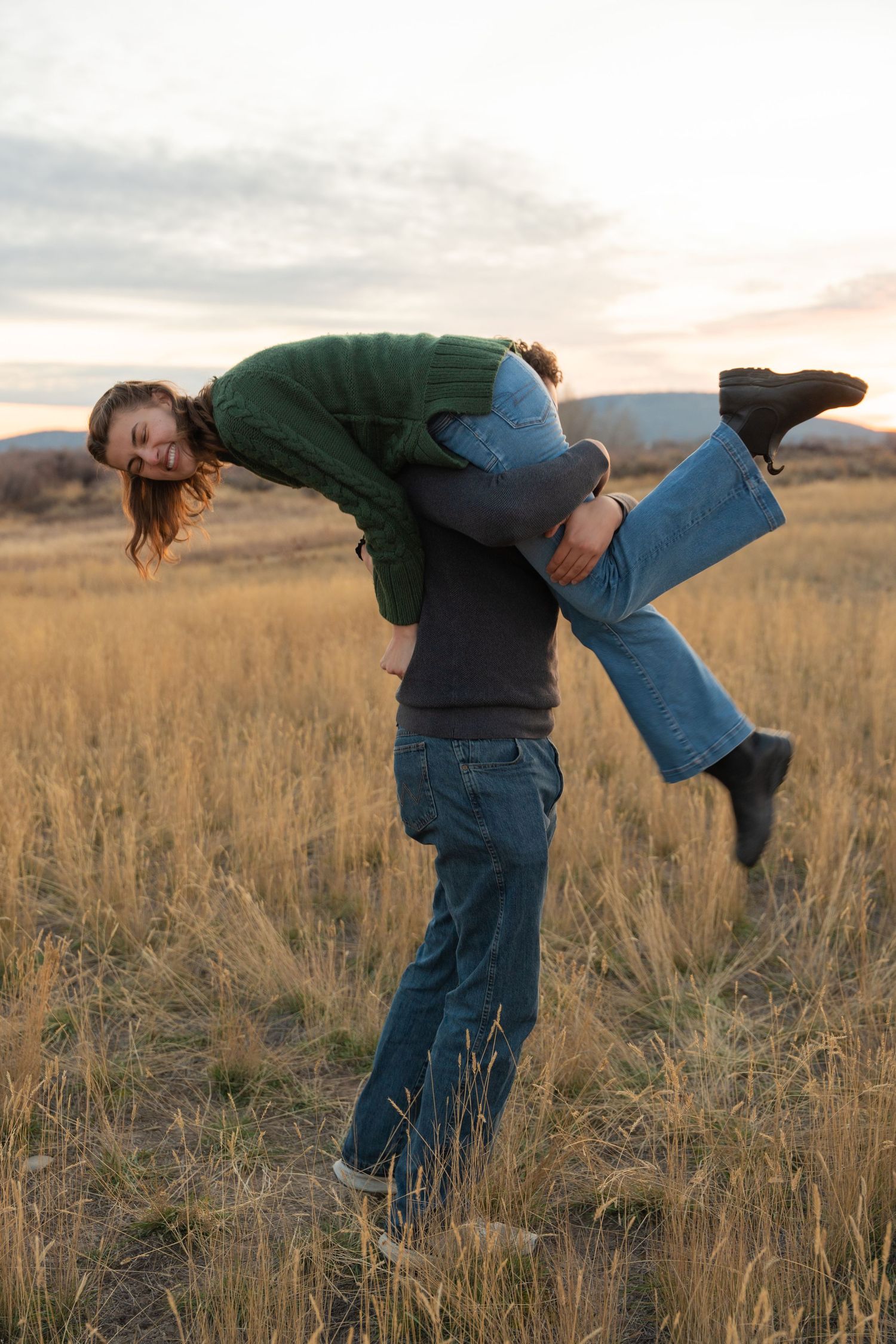 A playful couple embraces in a golden field of tall grass at sunset.