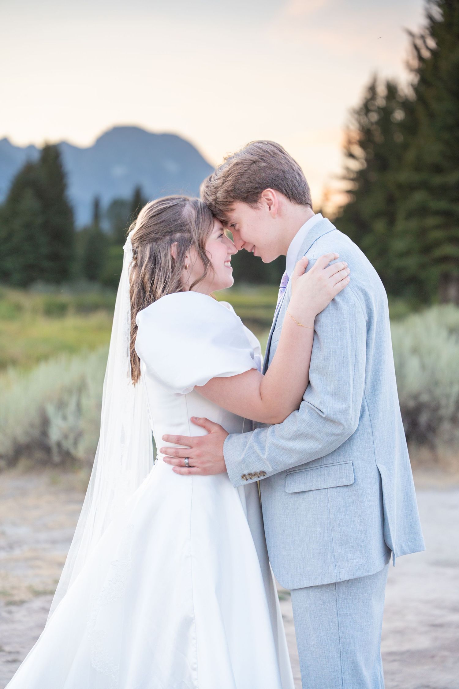 A romantic wedding couple embraces against a mountain backdrop at sunset.