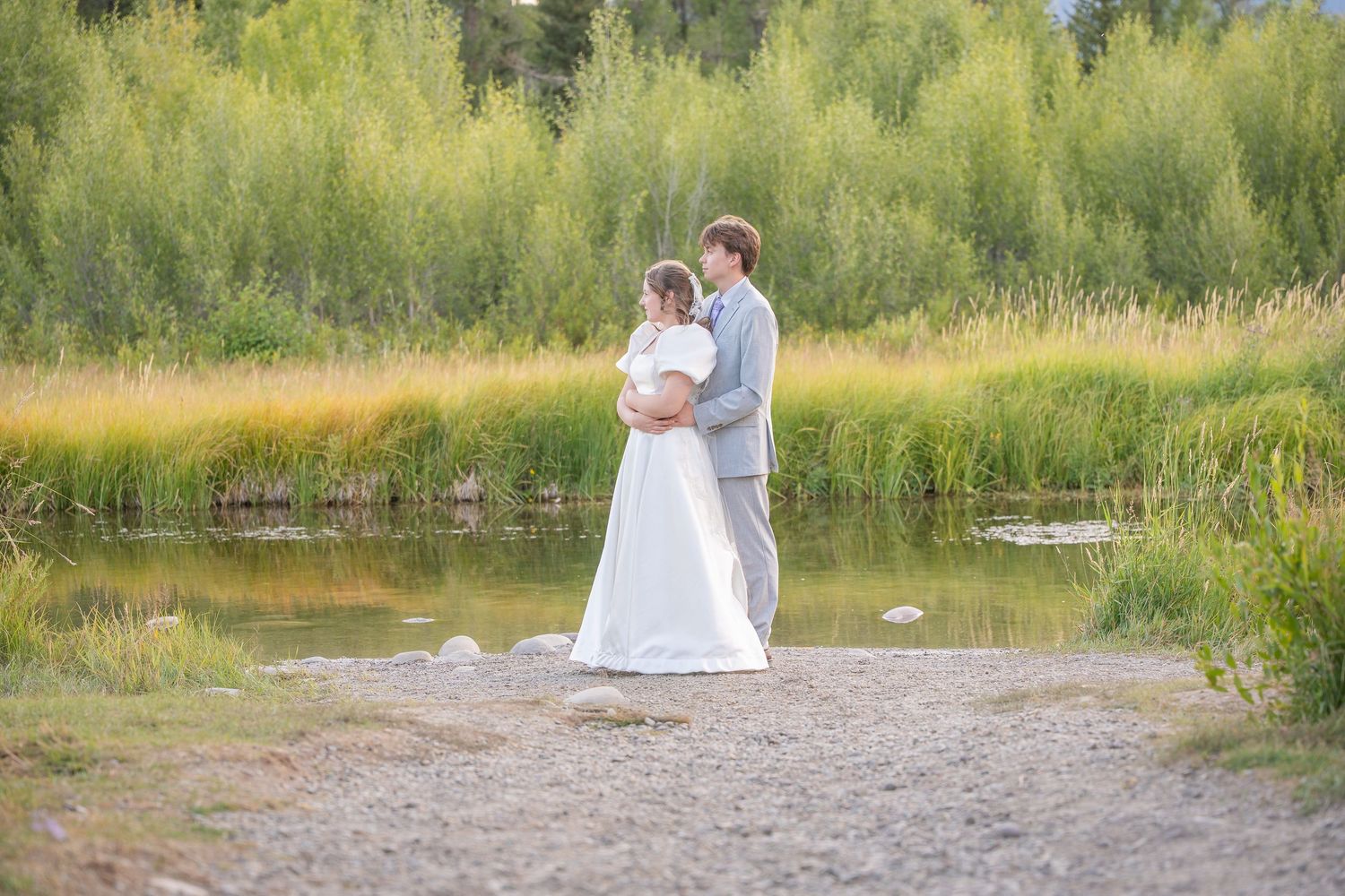 A serene wedding portrait of a couple standing by a lake with golden grasses and trees in the background.