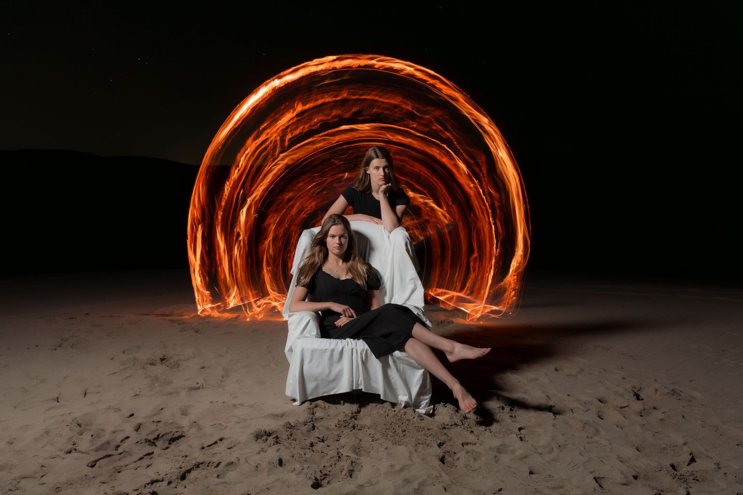 Long exposure light painting creates glowing orange spiral tunnel effect behind couple seated on beach at night.
