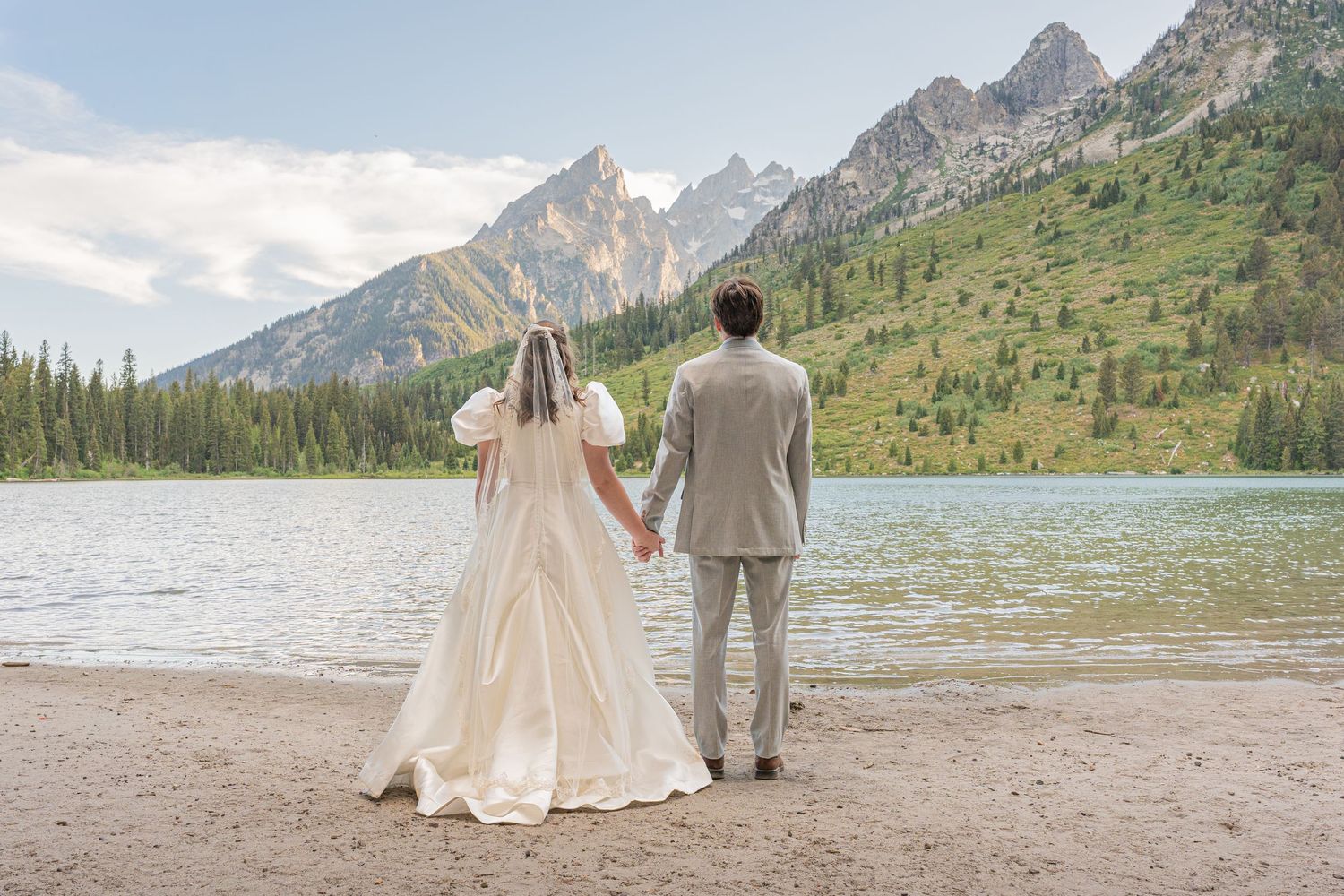 A couple in wedding attire holds hands while admiring a stunning mountain lake view.