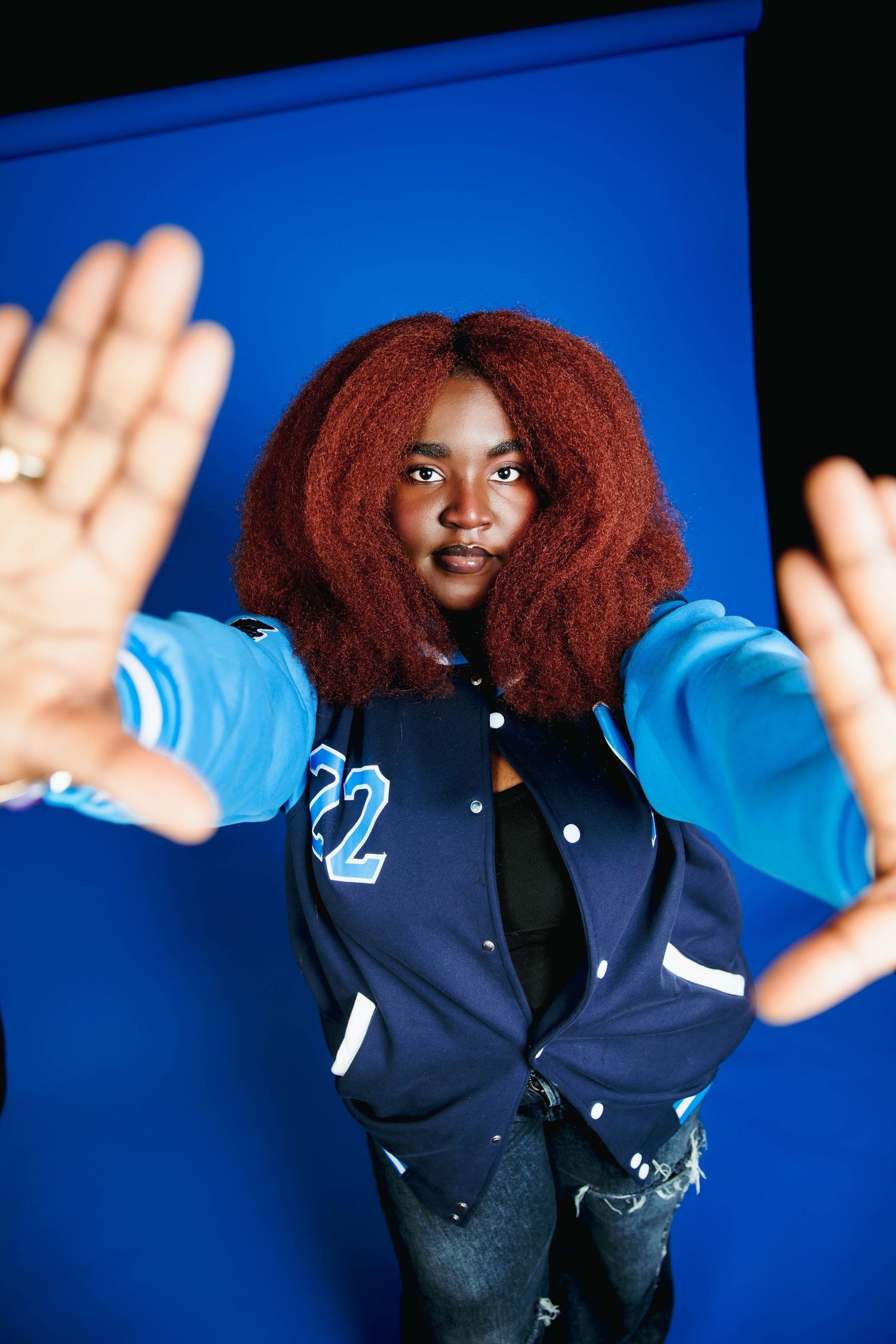 Young woman with red afro hair wearing a blue varsity jacket numbered 22, reaching hands toward camera against blue background.