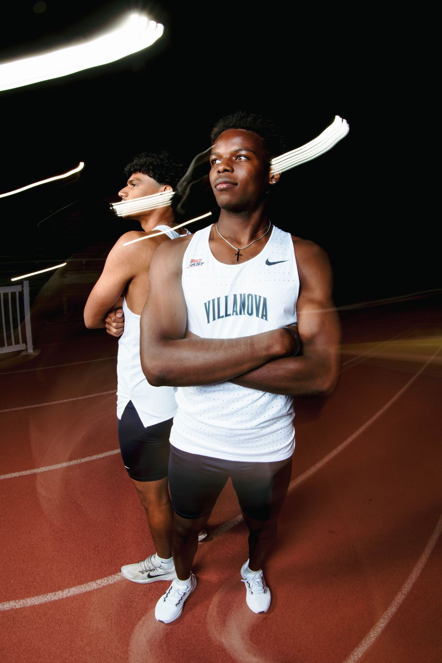 Two Villanova male track athletes in white Nike uniforms posing on red track with light trails at night.