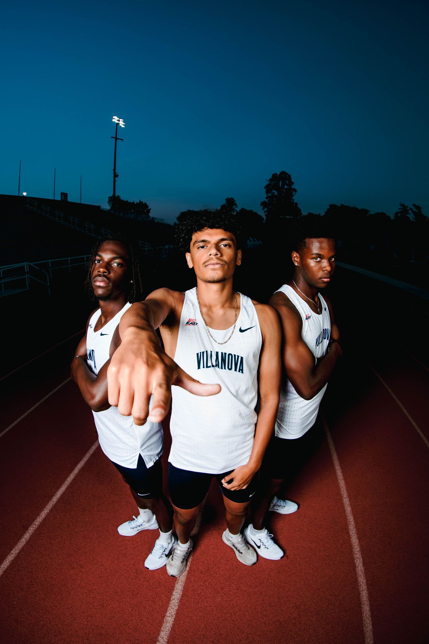 Three Villanova track athletes pose on a red track at dusk, center athlete pointing at camera.