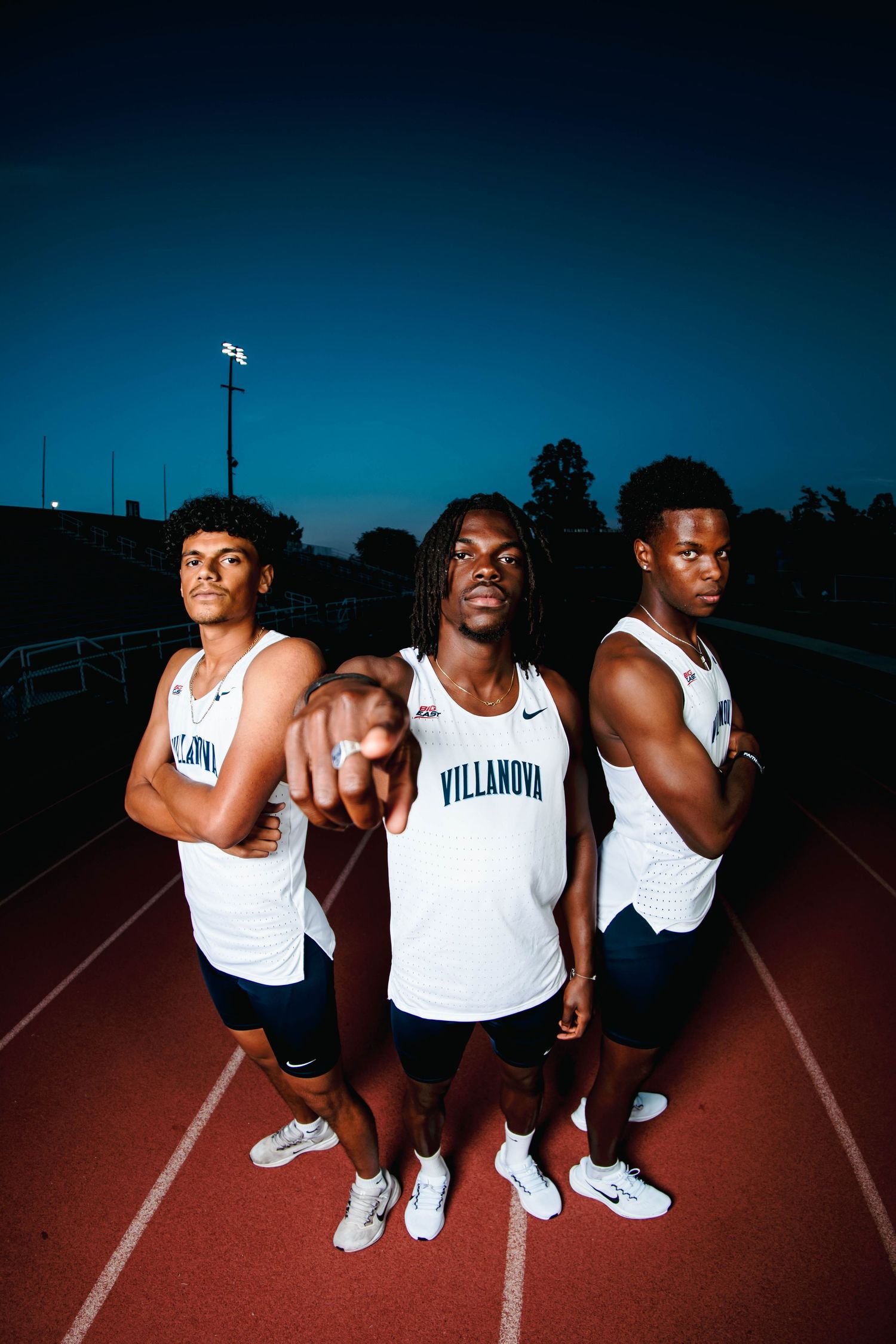 Three Villanova track athletes pose confidently on a red track at dusk in white uniforms.