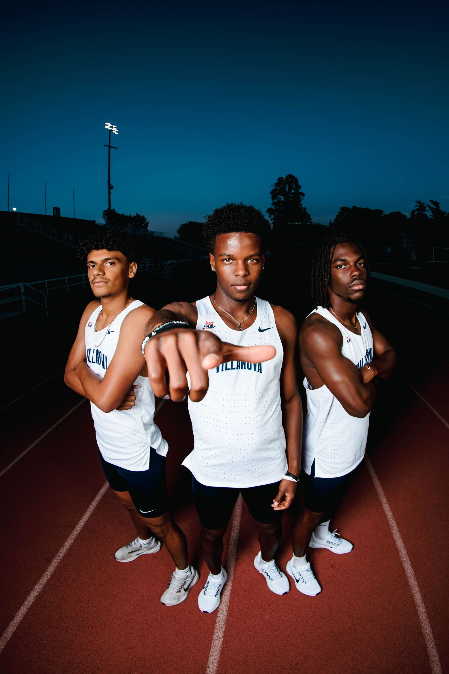 Three Villanova track and field athletes pose on a red track at dusk in white uniforms.