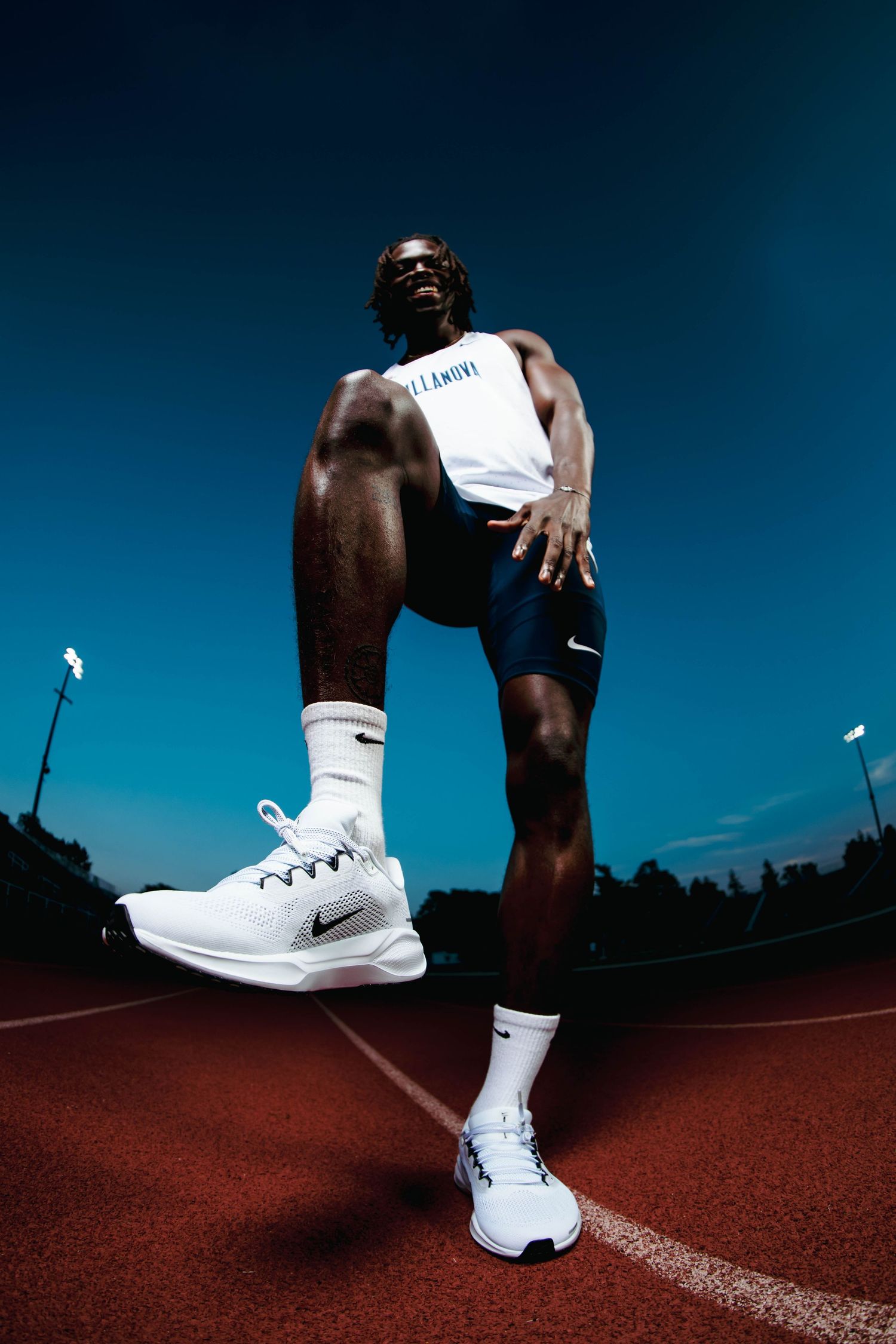 Male Villanova athlete in white Nike tank and shorts posing on red running track under blue sky.